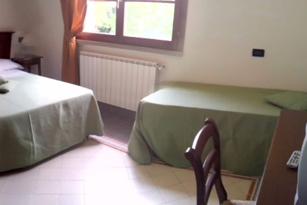 Quarto individual com uma cama de solteiro, cabeceira de madeira, mesa de cabeceira com abajur e janela com cortinas.
