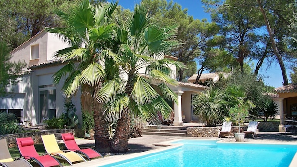 A swimming pool with lounge chairs, palm trees, and a building with a tiled roof. - Sous les Pins de Nages (Nages-et-Solorgues)