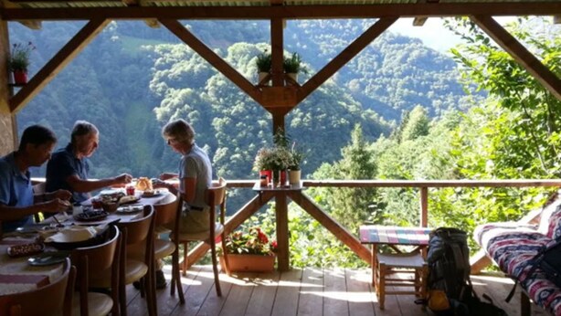 A wooden deck with a table and chairs, a sofa, and a view of a forested mountain range.
