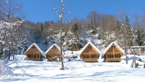 Four wooden cabins with snow-covered roofs in a snowy forest setting.