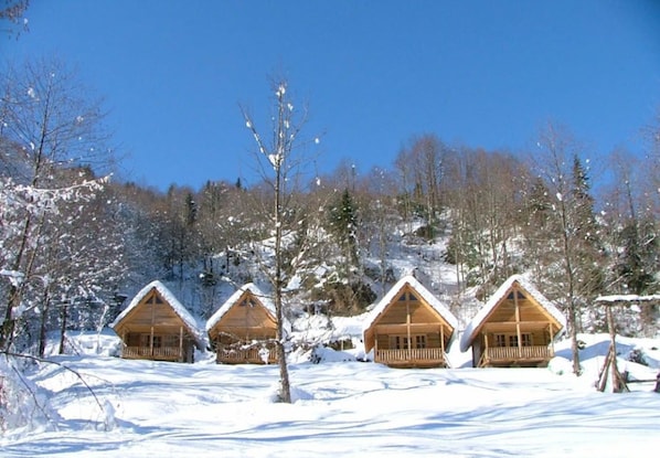 Four wooden cabins with snow-covered roofs in a snowy forest setting.