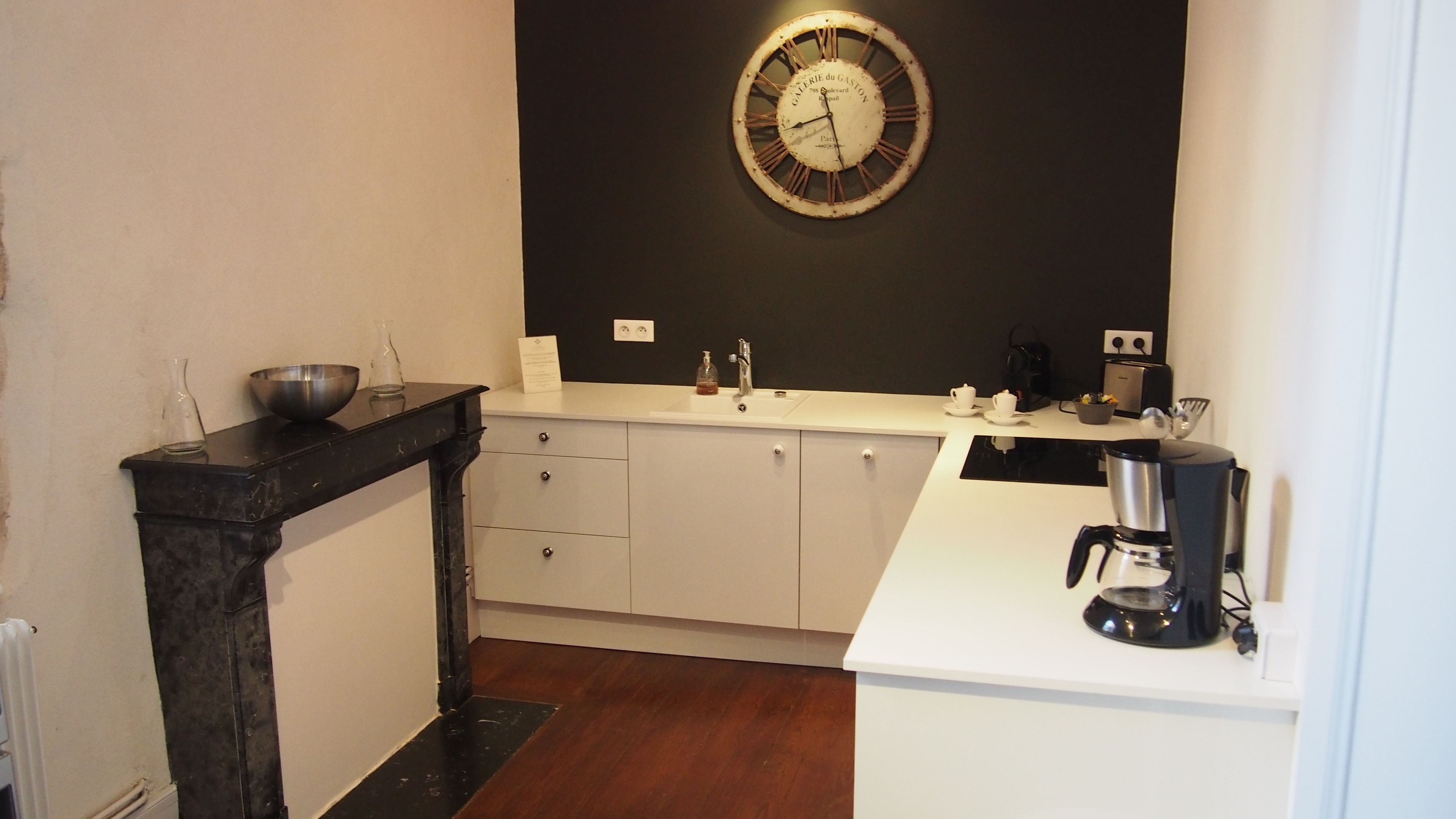 A kitchen with a black wall clock, white cabinets, a black countertop, and a coffee maker.