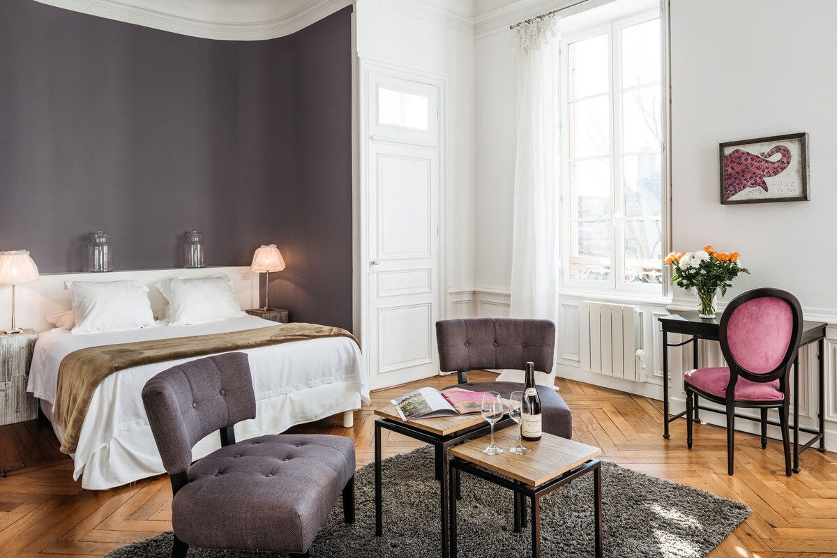 A bedroom with a bed, two chairs, a small table, a window, and a nightstand with a lamp.