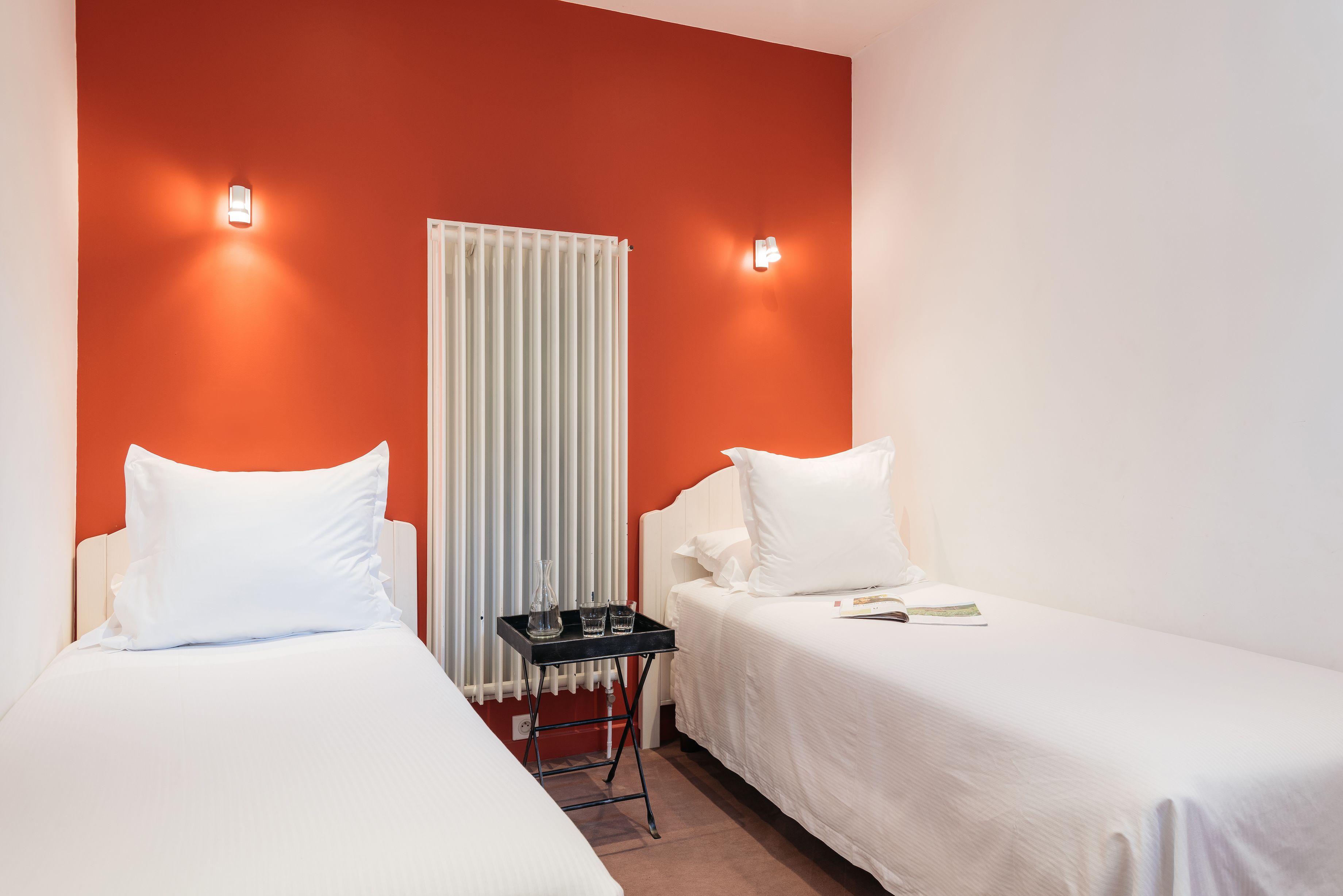 Une chambre d’hôtel avec deux lits simples, du linge de lit blanc, une table de chevet noire avec des verres et un radiateur.