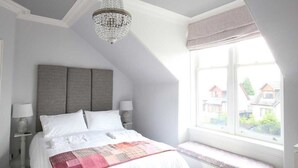 A bedroom with a large bed, a bench by the window, and a chandelier. - High Terrace Apartment (Grantown-on-Spey)