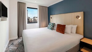 A hotel room with a large bed, a TV mounted on the wall, and a window with curtains. - Sydney Suites (Sydney)