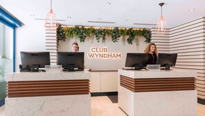 A hotel reception area with two reception desks labeled "HOTEL RECEPTION" and "INFORMATION DESK," both with computer monitors. The reception area is decorated with green plants and has a sign that reads "CLUB WYNDHAM." - Sydney Suites (Sydney)