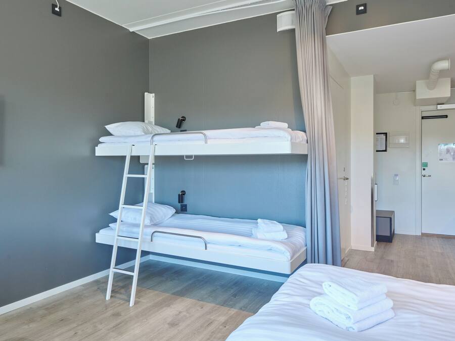Una habitación de hotel con literas, una mesita de noche, una lámpara y una puerta que da a un baño.