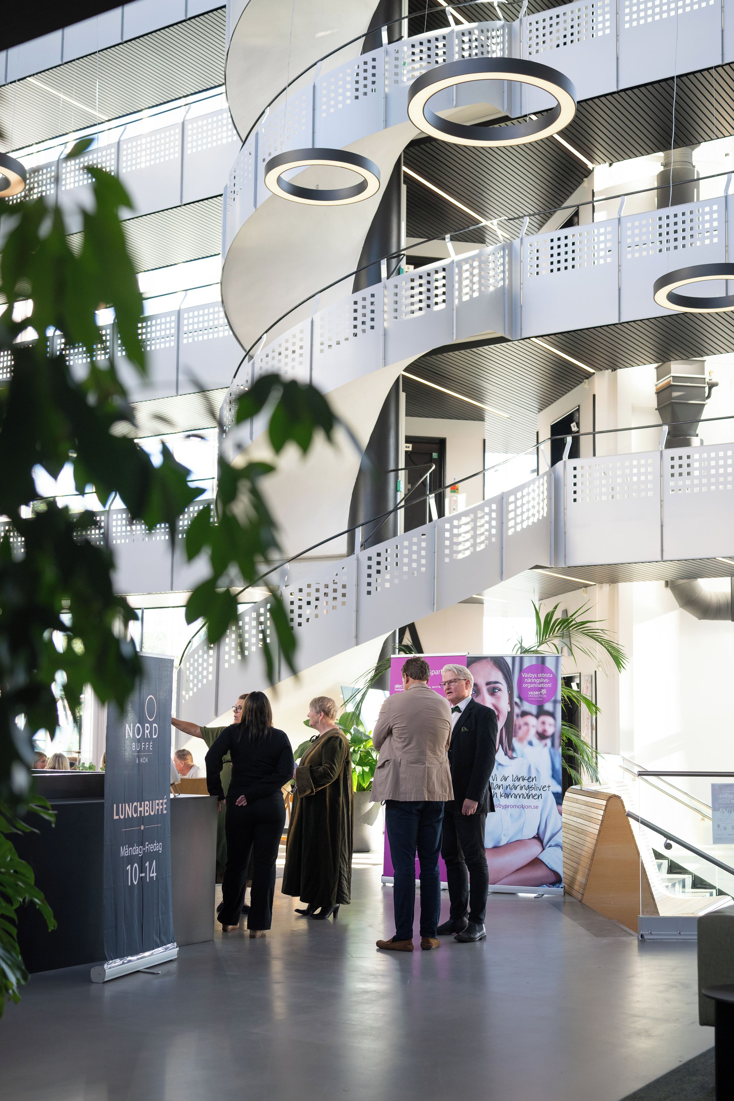 A modern interior with a central staircase, a sign for a lunch buffet, and people standing around.