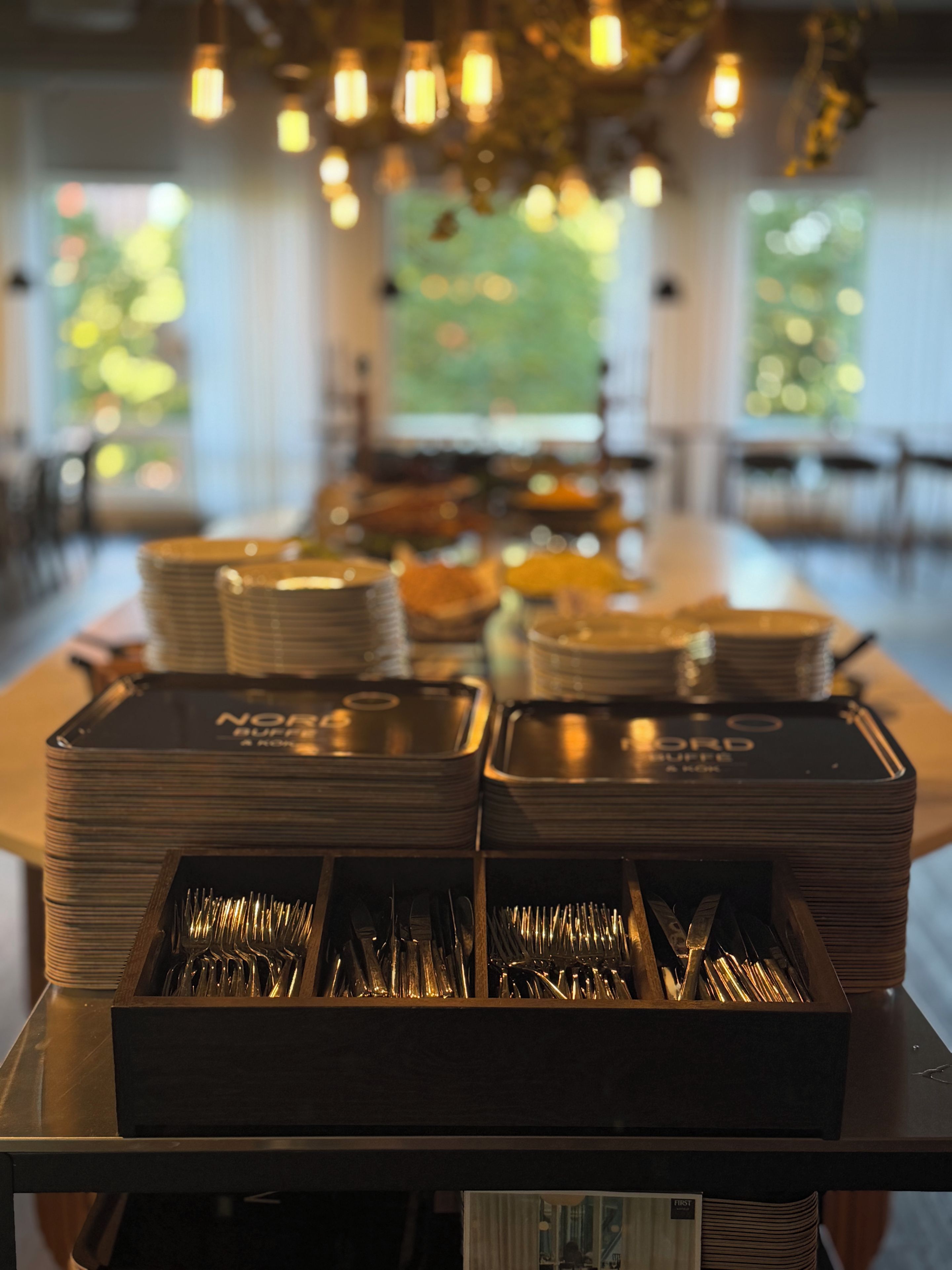 Montaje tipo buffet con platos apilados y cubiertos en cajas de madera, con luces colgantes en el techo.