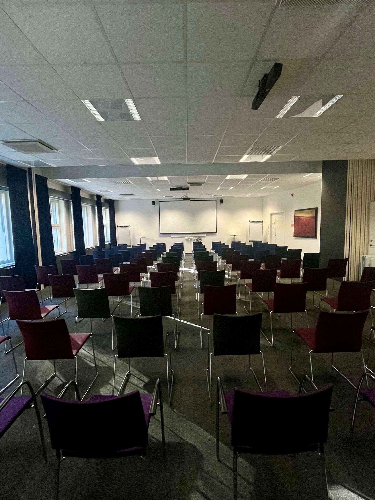 Ein Konferenzraum mit Reihen von Stühlen, einer Projektionsleinwand und Fenstern.