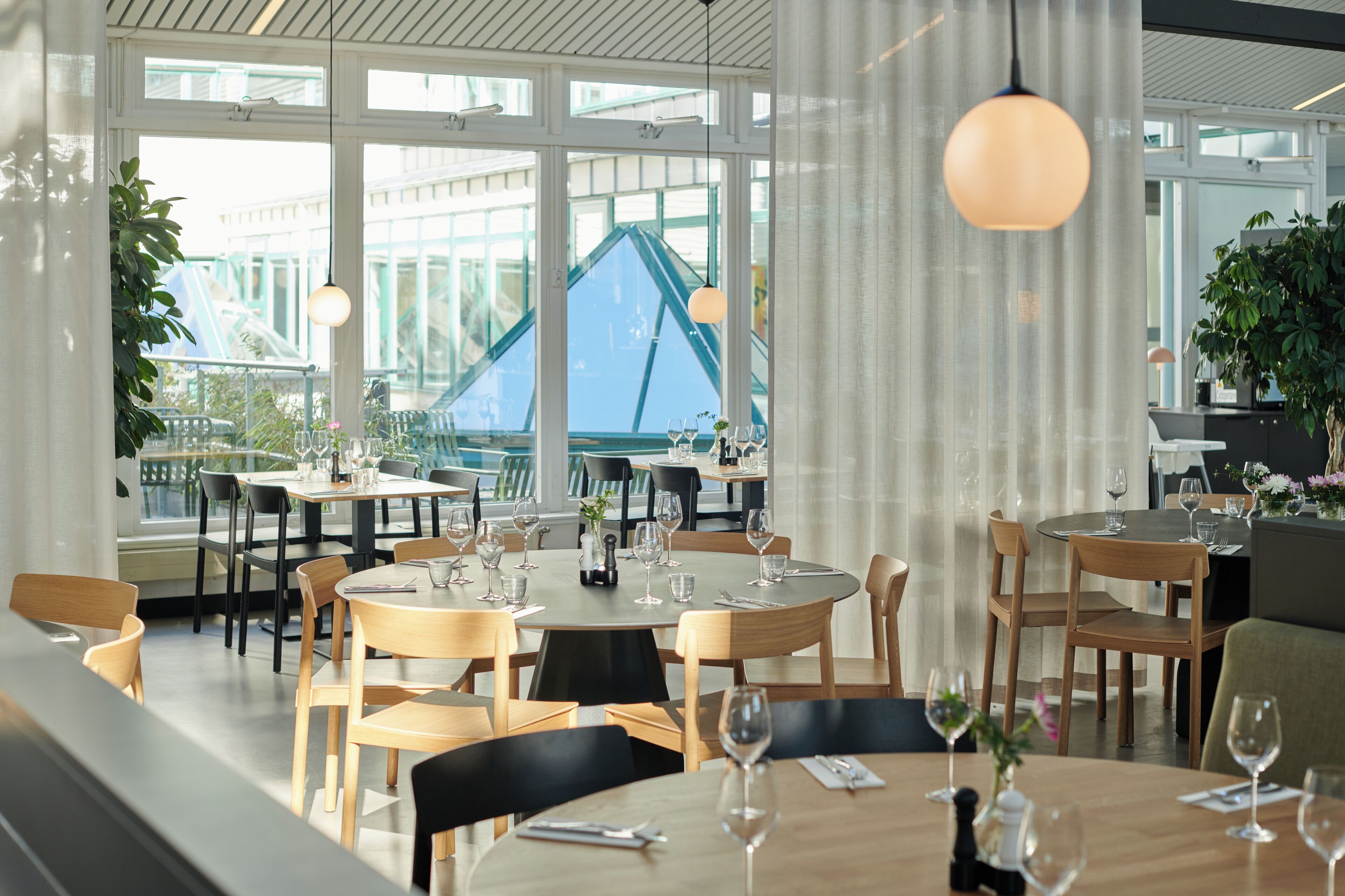 Un restaurante con mesas y sillas redondas, ventanas amplias y lámparas colgantes.