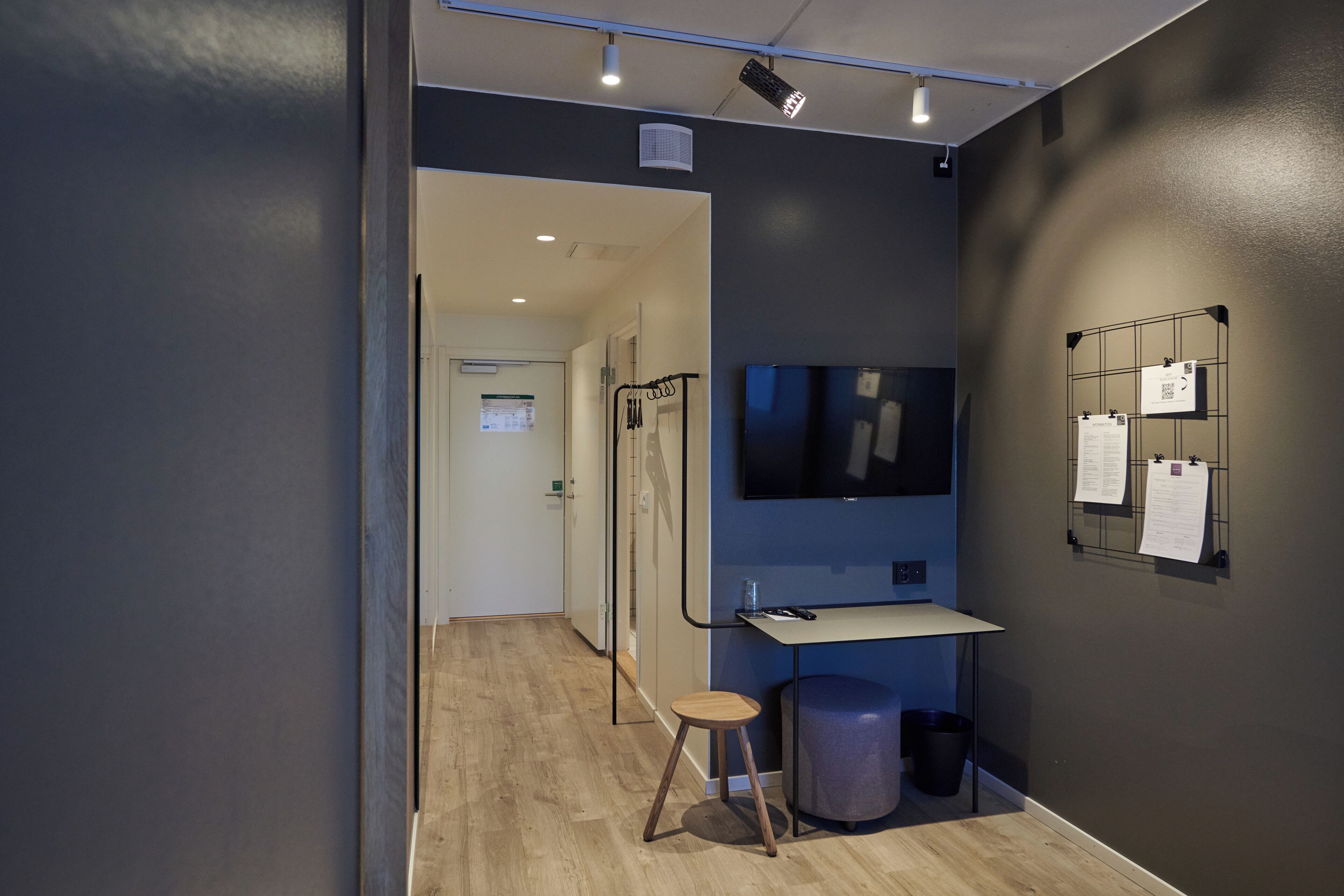 A modern room with a desk, chair, and a mounted TV. There are papers on the wall and a trash bin.