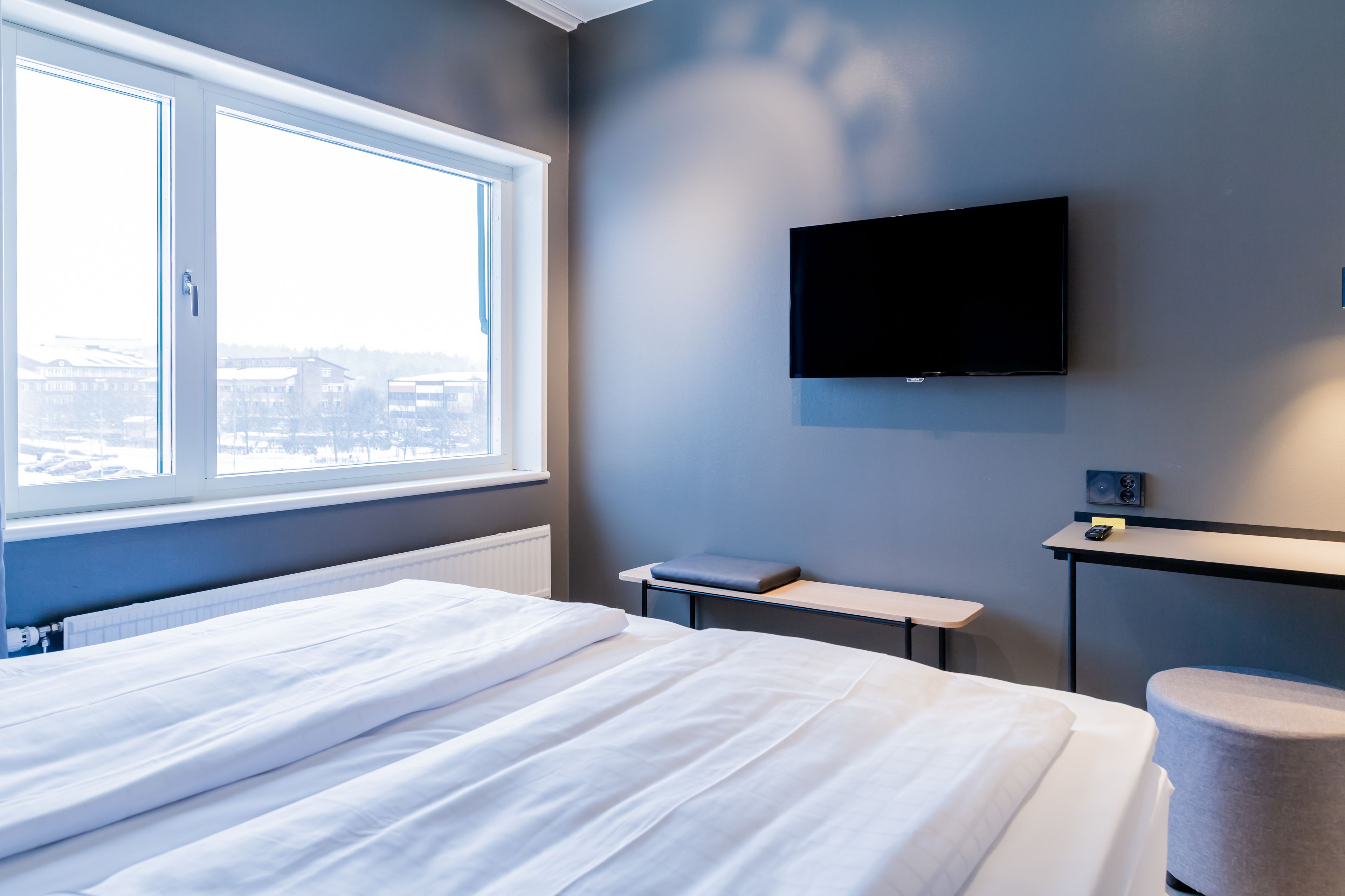 A hotel room with a bed, a desk, a chair, a TV, and a window with a view of buildings.