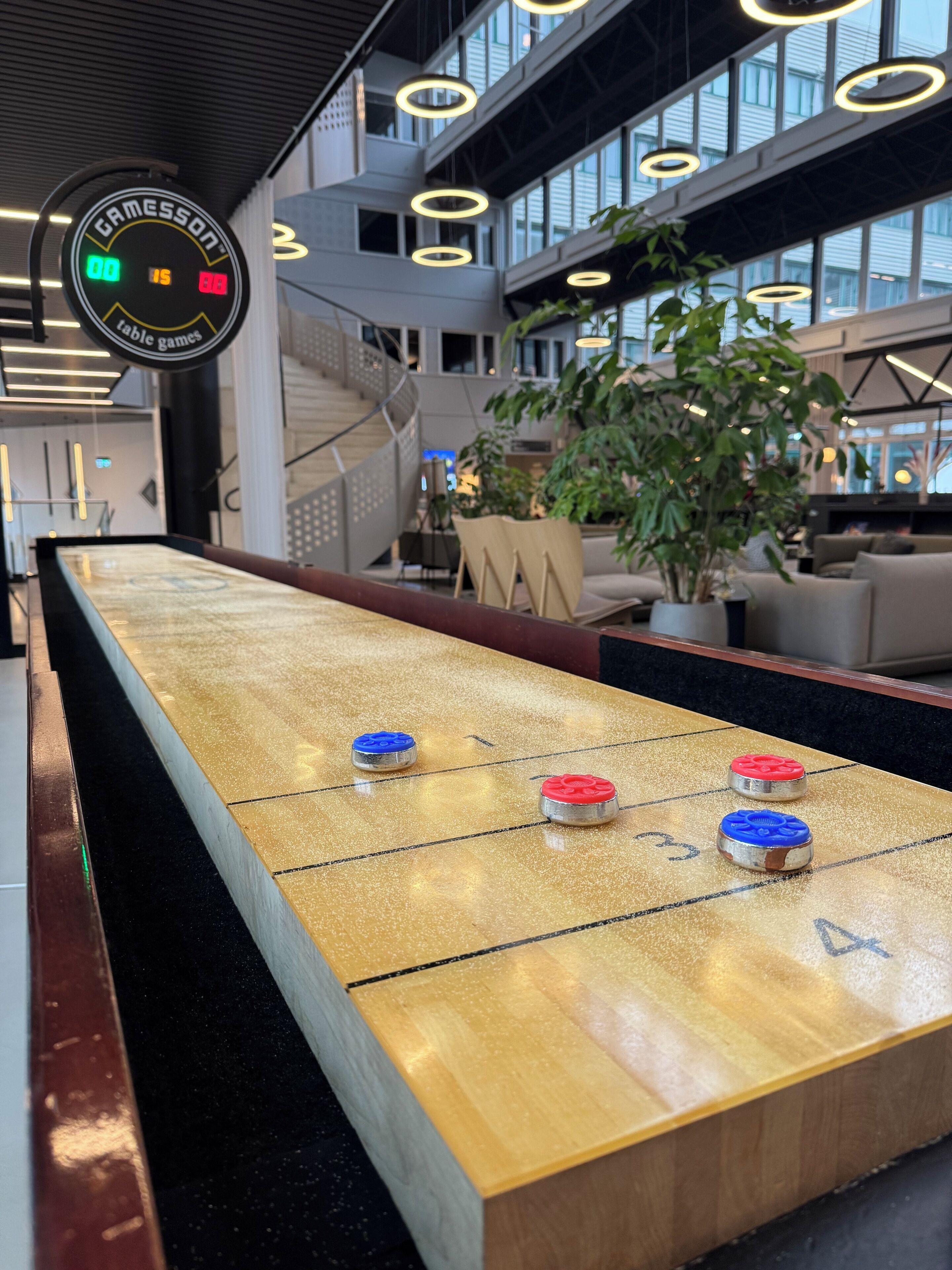A shuffleboard table with pucks, a digital scoreboard, and a modern interior with large windows and plants.