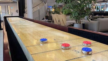 A shuffleboard table with pucks, a digital scoreboard, and a modern interior with large windows and plants.