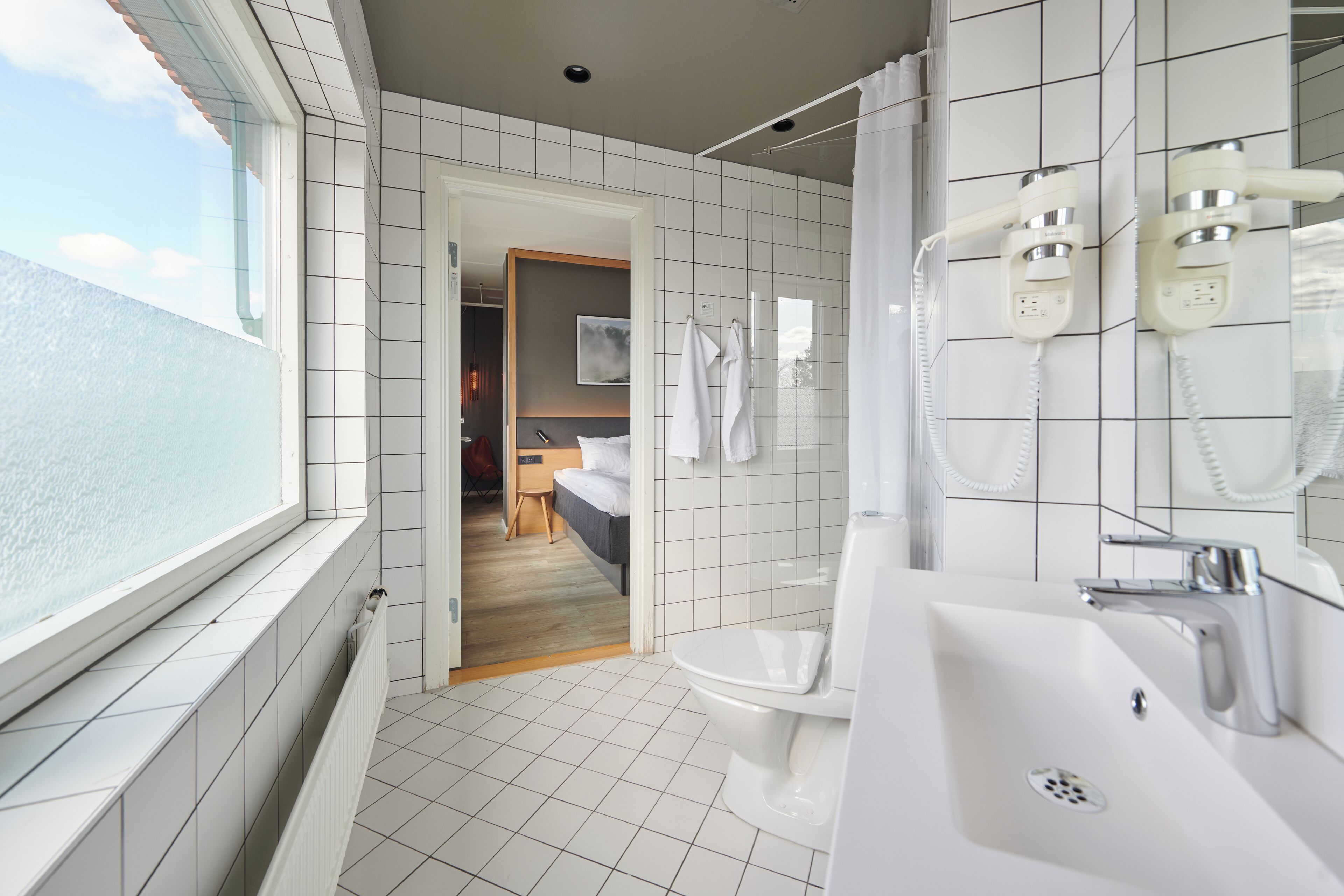 Un baño moderno con azulejos blancos, un ventanal grande, una ducha con cabezal de ducha manual y un lavamanos con espejo.