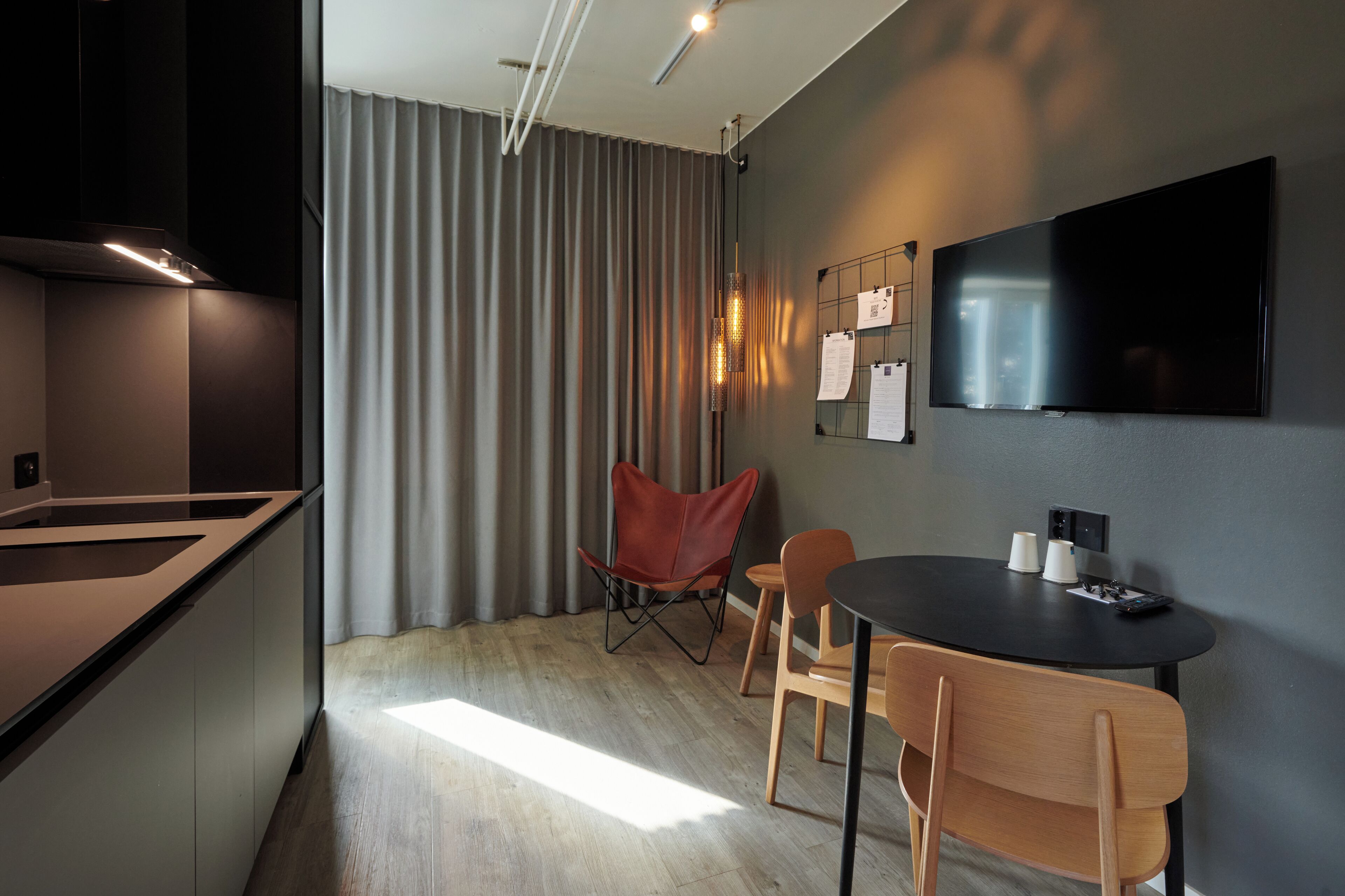 A modern kitchen with a dining area, a flat-screen TV mounted on the wall, and a window with curtains.