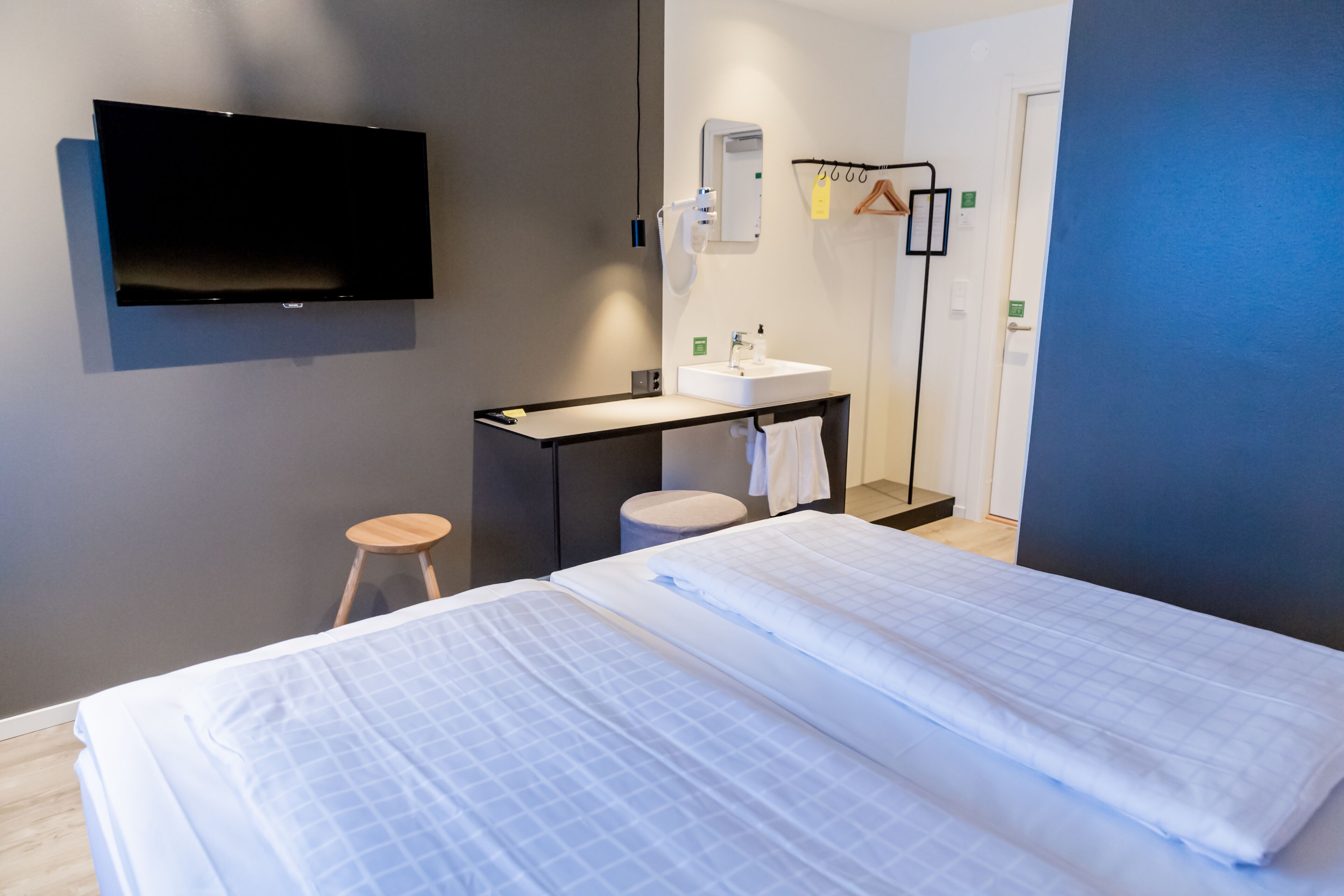 A hotel room with two beds, a flat-screen TV mounted on the wall, a small desk with a sink, and a mirror.