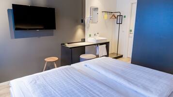 A hotel room with two beds, a flat-screen TV mounted on the wall, a small desk with a sink, and a mirror.