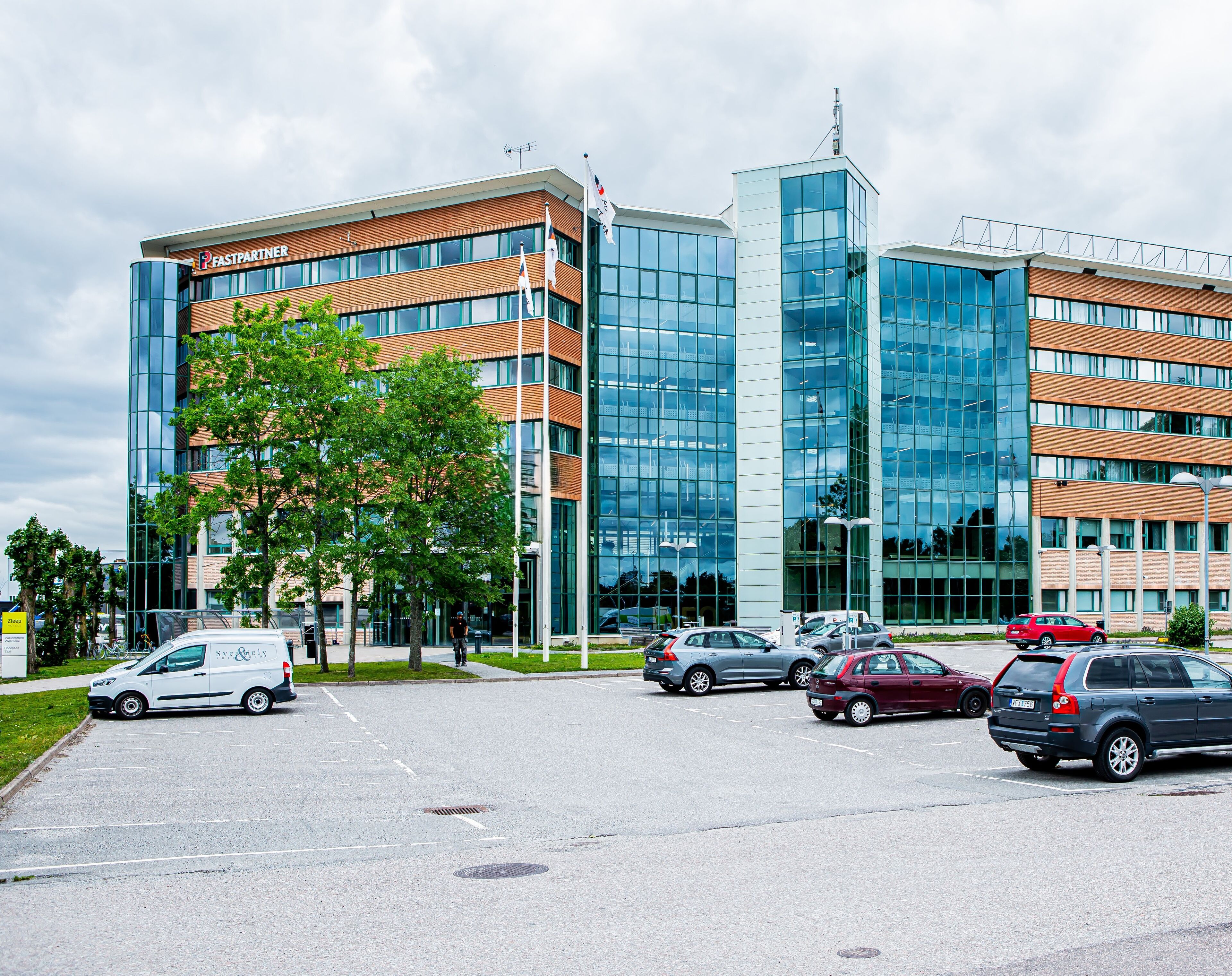 Un bâtiment moderne à plusieurs étages, doté de grandes fenêtres et d’un parking où sont stationnées plusieurs voitures.