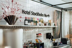 Zona de bar con una variedad de botellas y vasos, un ventilador con detalles decorativos rojos y un letrero que dice 'Hotel Stockholm North'.