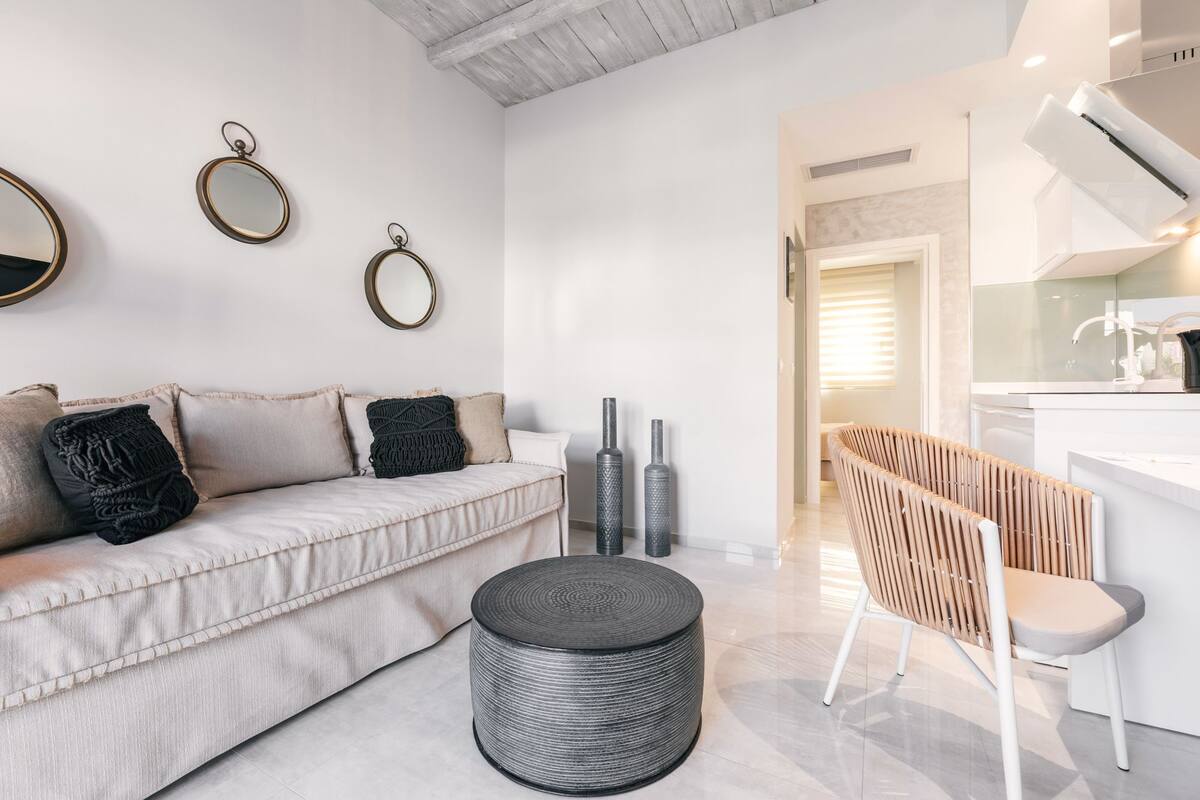 Uma sala de estar moderna com um sofá, um banquinho redondo e uma área de jantar com uma cadeira e uma mesa.