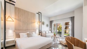 A modern hotel room with a large bed, a sofa, a chair, and a balcony with a view of the garden.