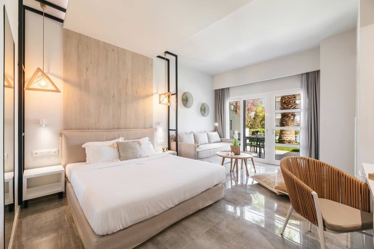 Quarto de hotel moderno com uma cama grande, um sofá, uma cadeira e uma varanda com vista para o jardim.