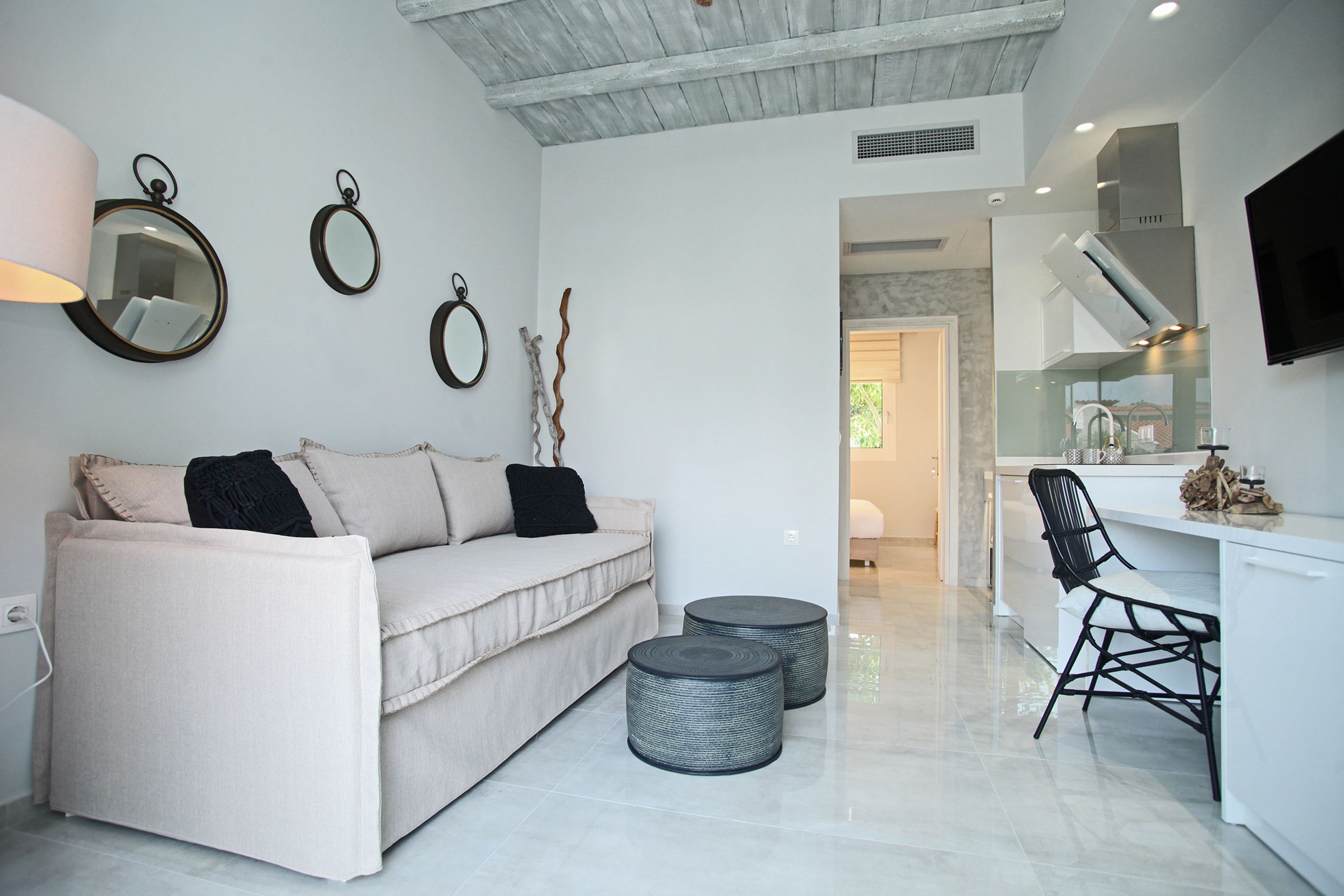 A modern living room with a sofa, two round ottomans, a black chair, and a kitchen area with a TV.