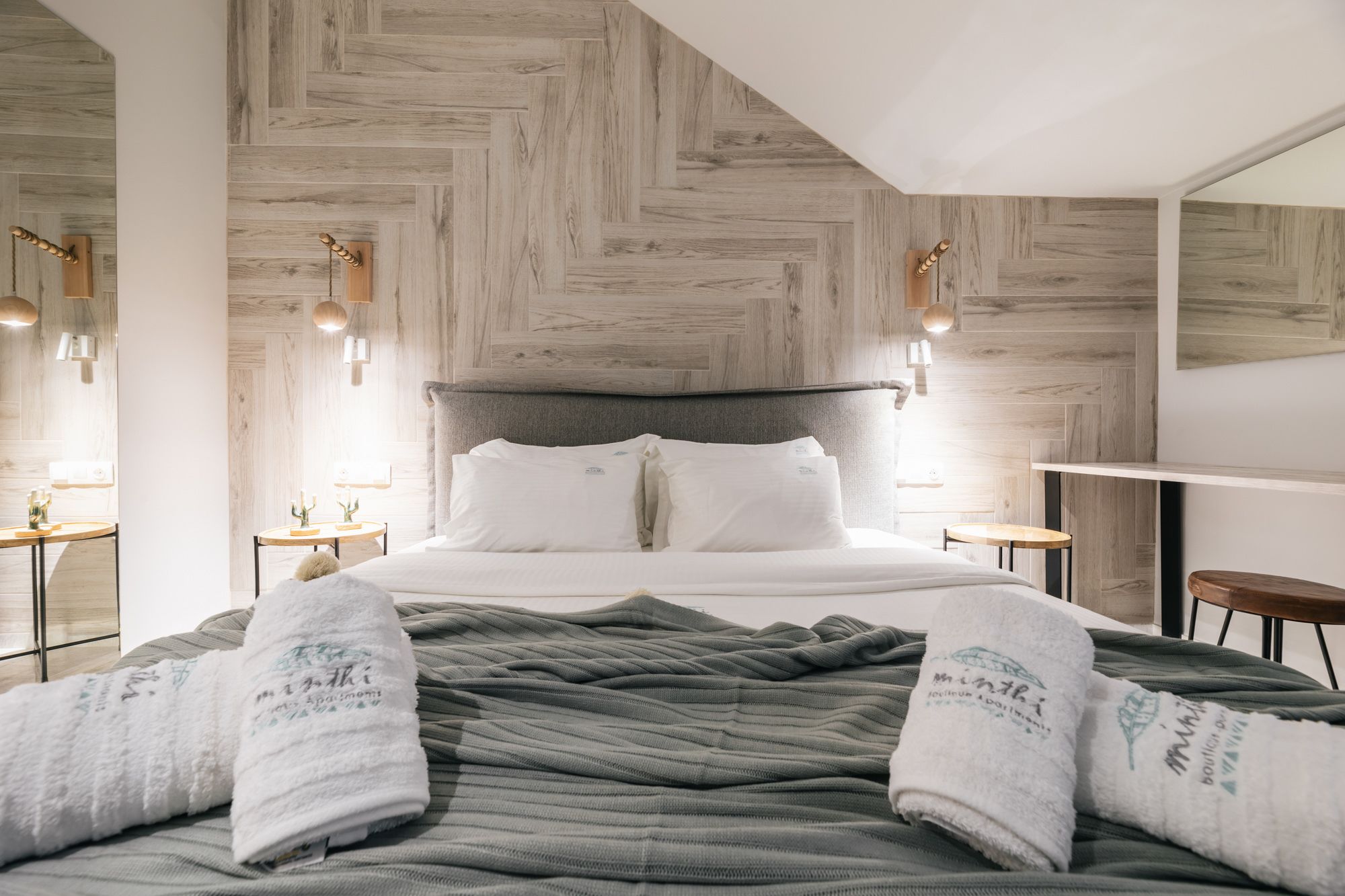 Une chambre d’hôtel moderne avec un mur en bois à motifs en chevrons, un grand lit avec du linge de lit blanc, deux tables de chevet avec des lampes et une petite table d’appoint en bois.
