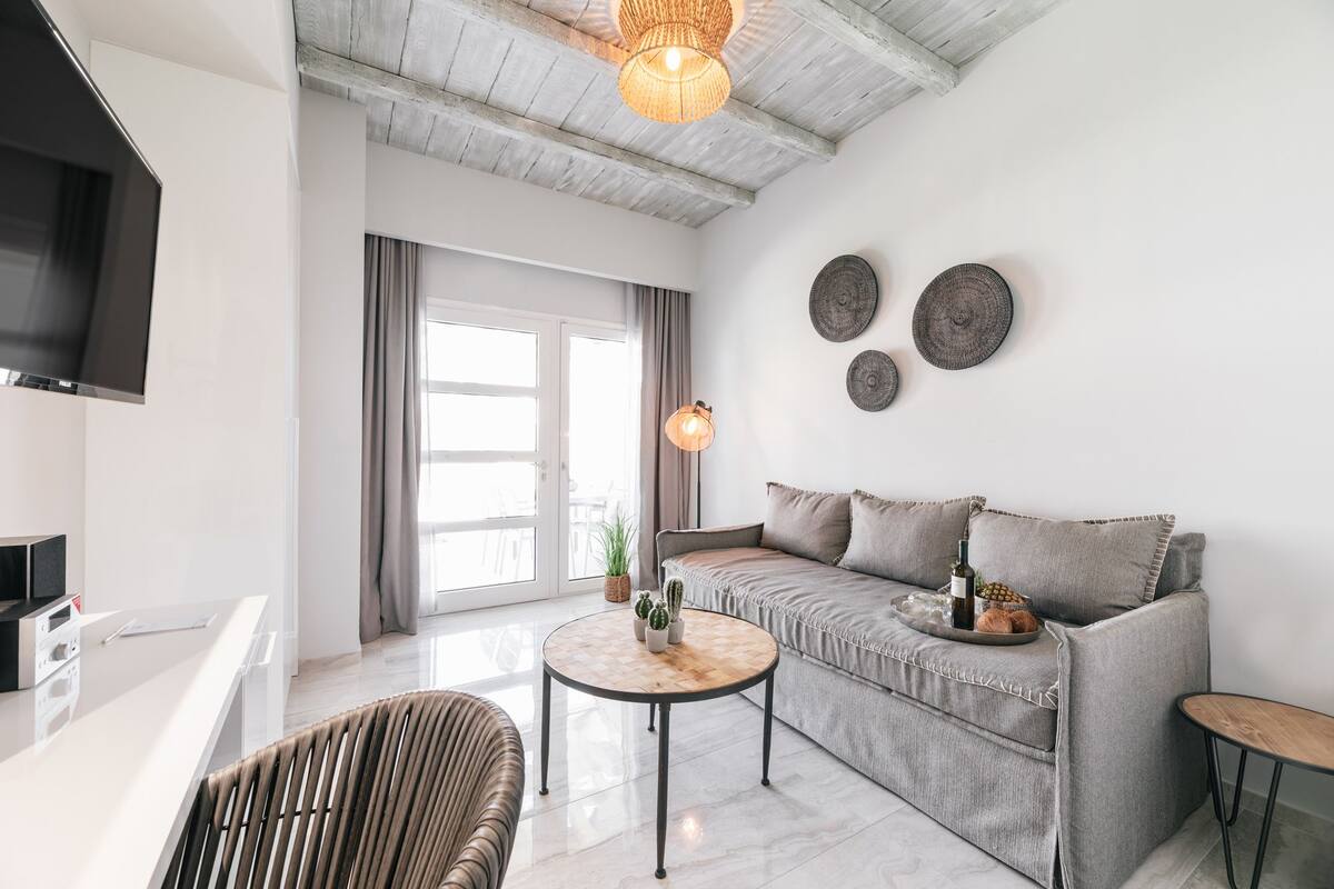 Uma sala de estar moderna com um sofá cinzento, uma mesa de centro redonda de madeira, uma poltrona de vime e uma TV de tela plana na parede.