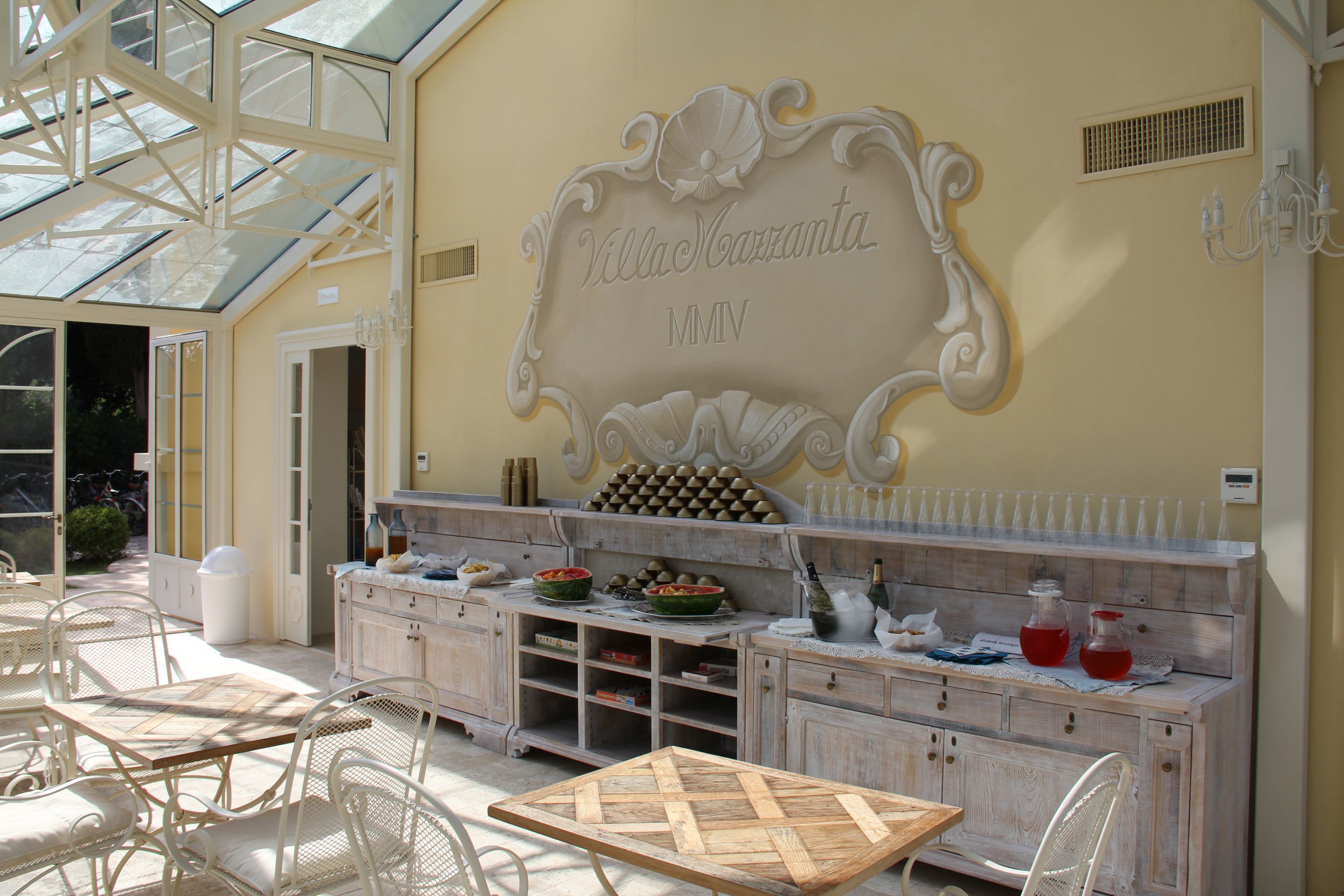 Un patio soleado con una mesa y sillas de madera, una pared decorativa con el texto 'Villa Maranta MMIV' y una estantería para vinos.