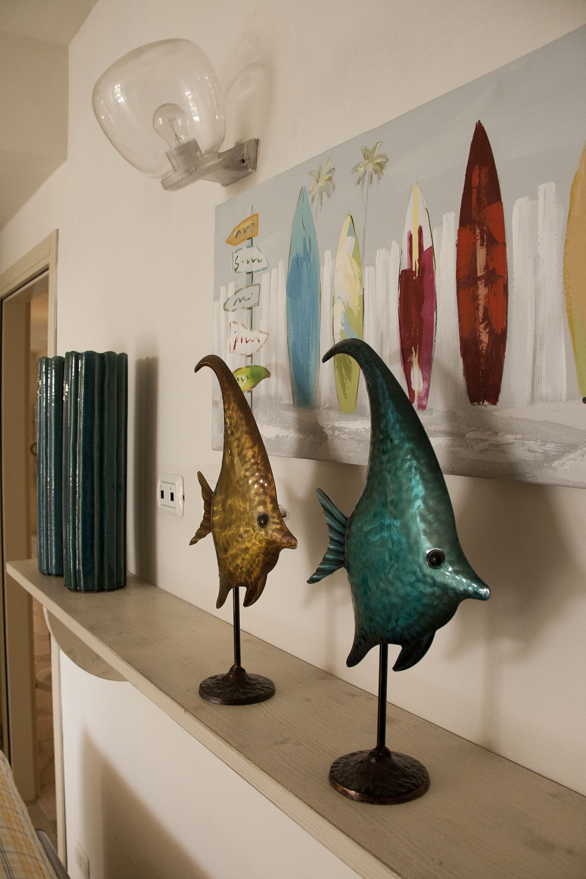 A hallway with a wooden shelf displaying two fish sculptures, a wall-mounted light fixture, and a colorful painting of surfboards.