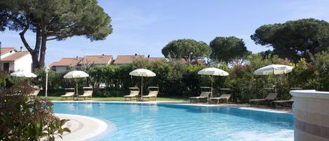 Piscina con sillas de descanso y sombrillas, rodeada de árboles y edificios.