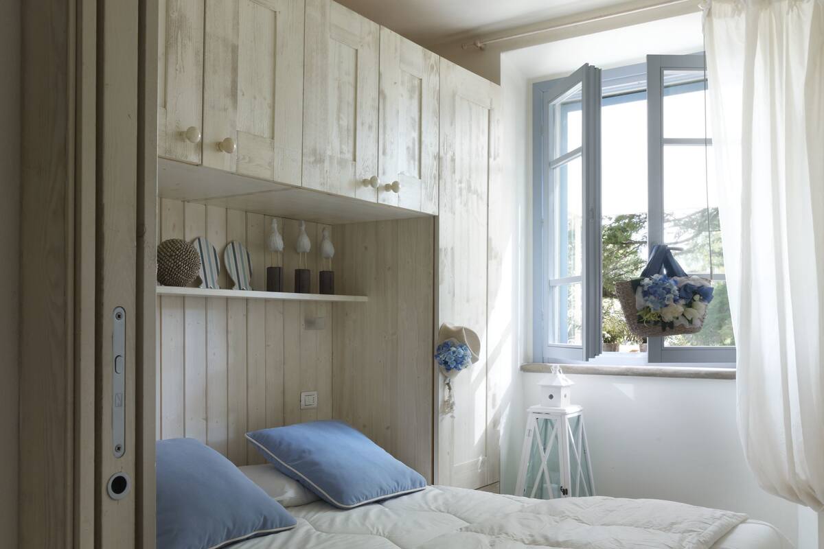 A bedroom with a bed, a window with curtains, a shelf with decorative items, and a door.