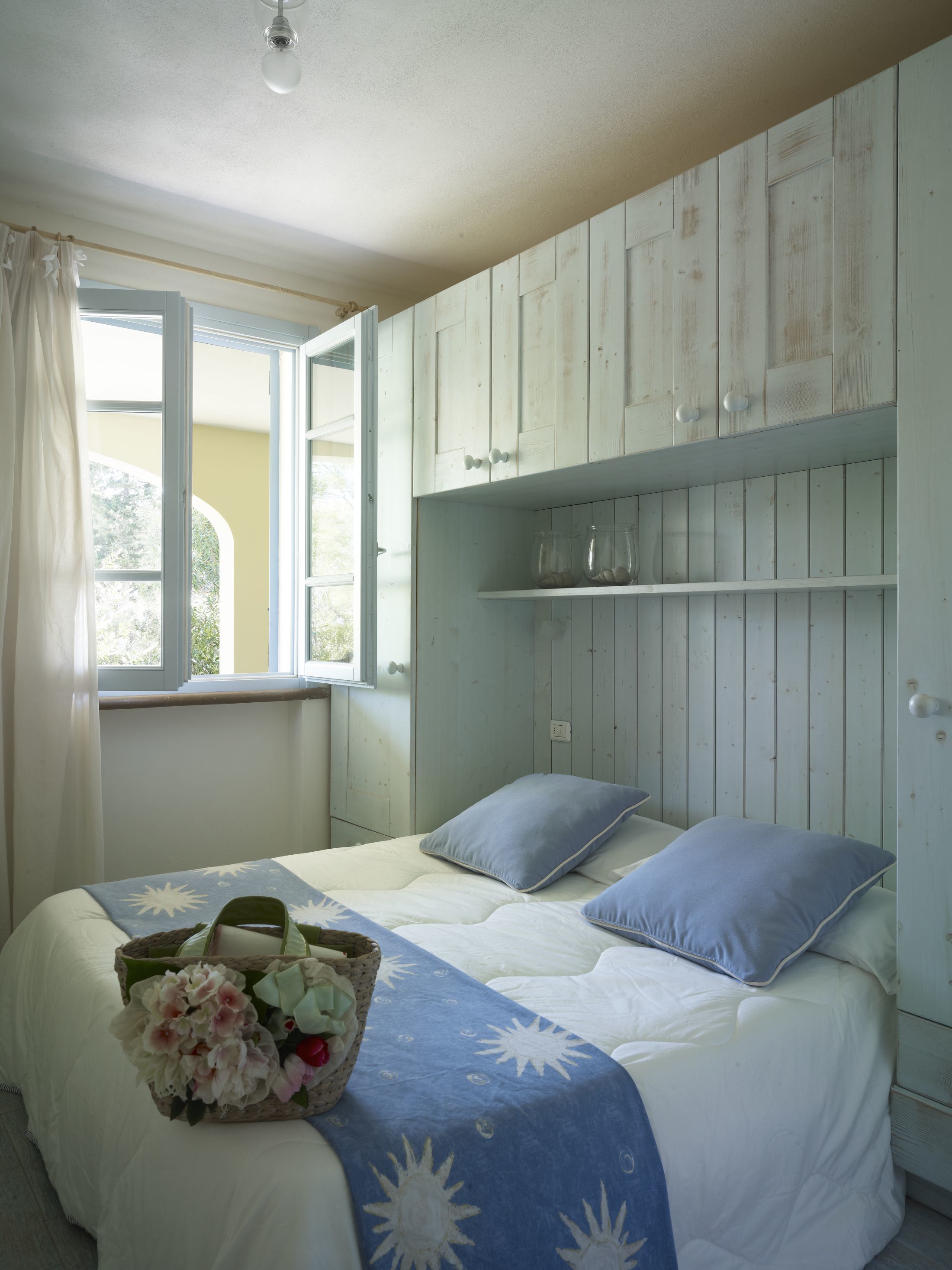 Een slaapkamer met een bed, kussens, een bloemenmand, een raam met gordijnen en houten lambrisering.