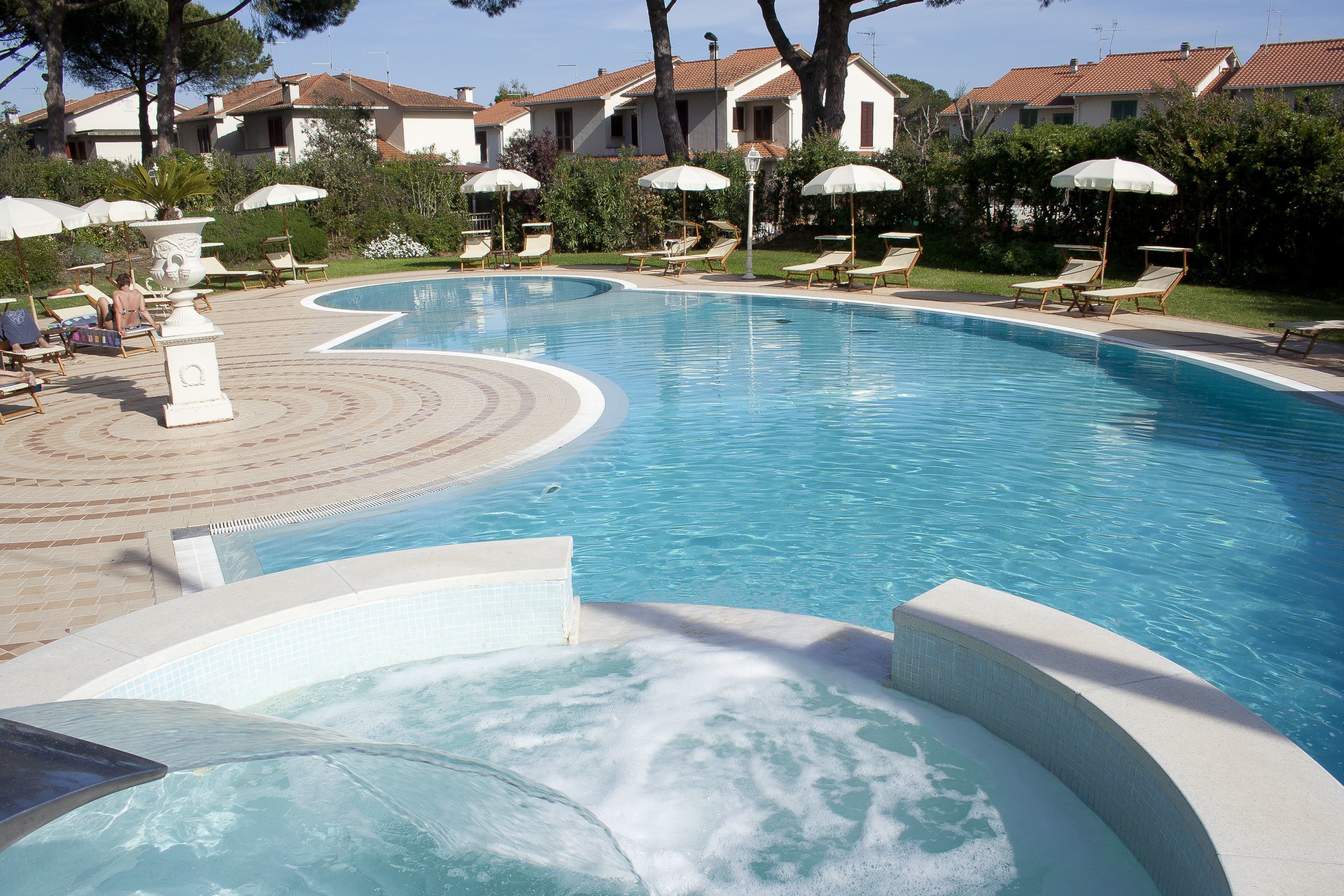 Ein Swimmingpool mit Liegestühlen und Sonnenschirmen, umgeben von einem angelegten Garten und Gebäuden im Hintergrund.