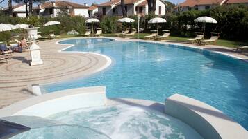 Ein Swimmingpool mit Liegestühlen und Sonnenschirmen, umgeben von einem angelegten Garten und Gebäuden im Hintergrund.