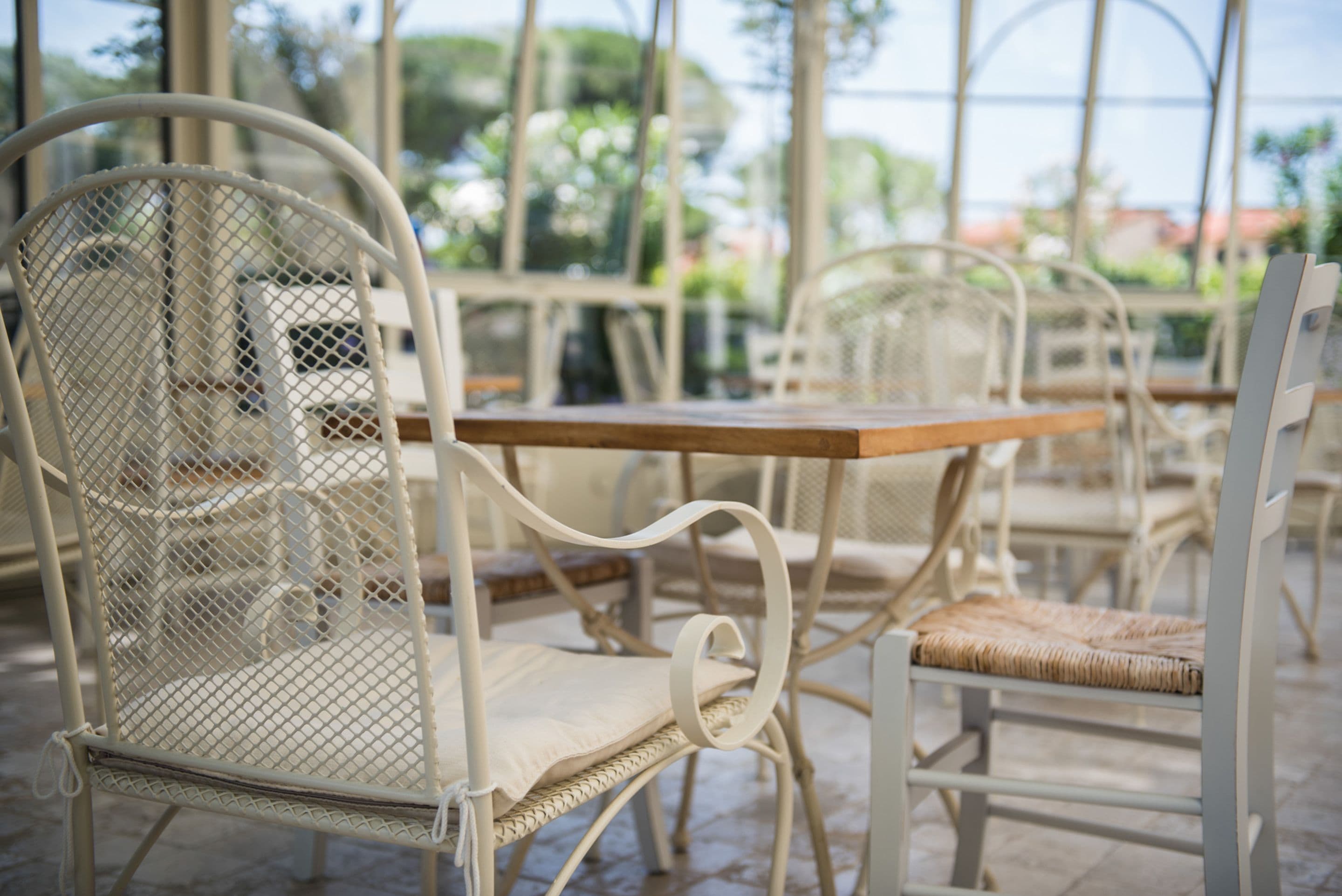 Una silla metálica blanca con asiento y respaldo acolchados, ubicada frente a una mesa de madera en un espacio exterior con techo de vidrio y follaje al fondo.