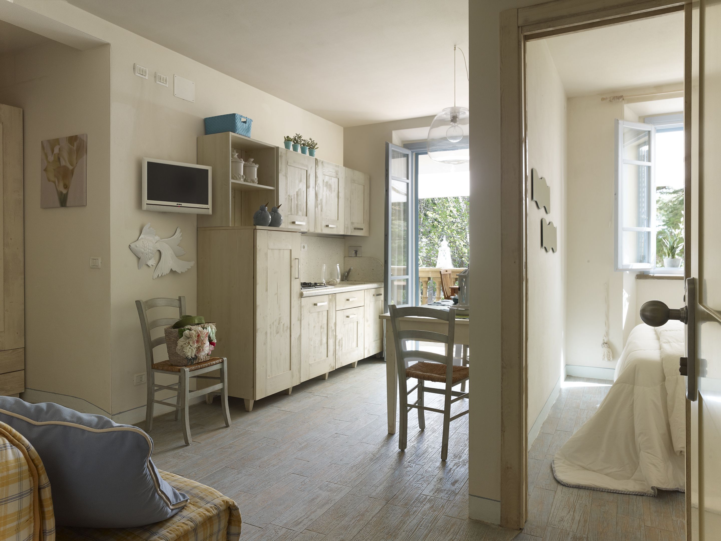 A cozy kitchen with wooden cabinets, a dining table, and chairs. There is a TV mounted on the wall, and a window providing natural light.