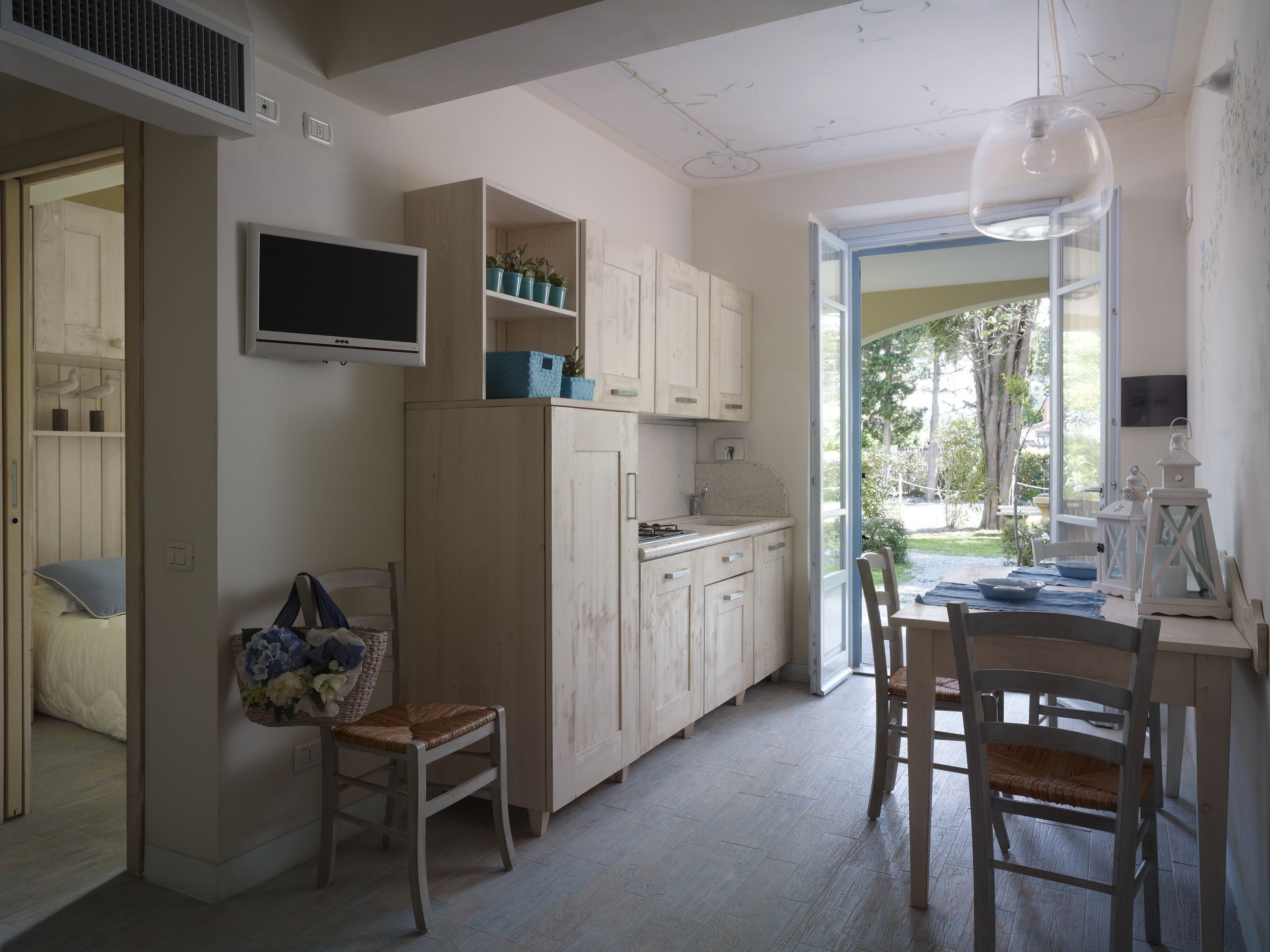 A kitchen with wooden cabinets, a dining table with chairs, a TV mounted on the wall, and a window providing a view of the outdoors.