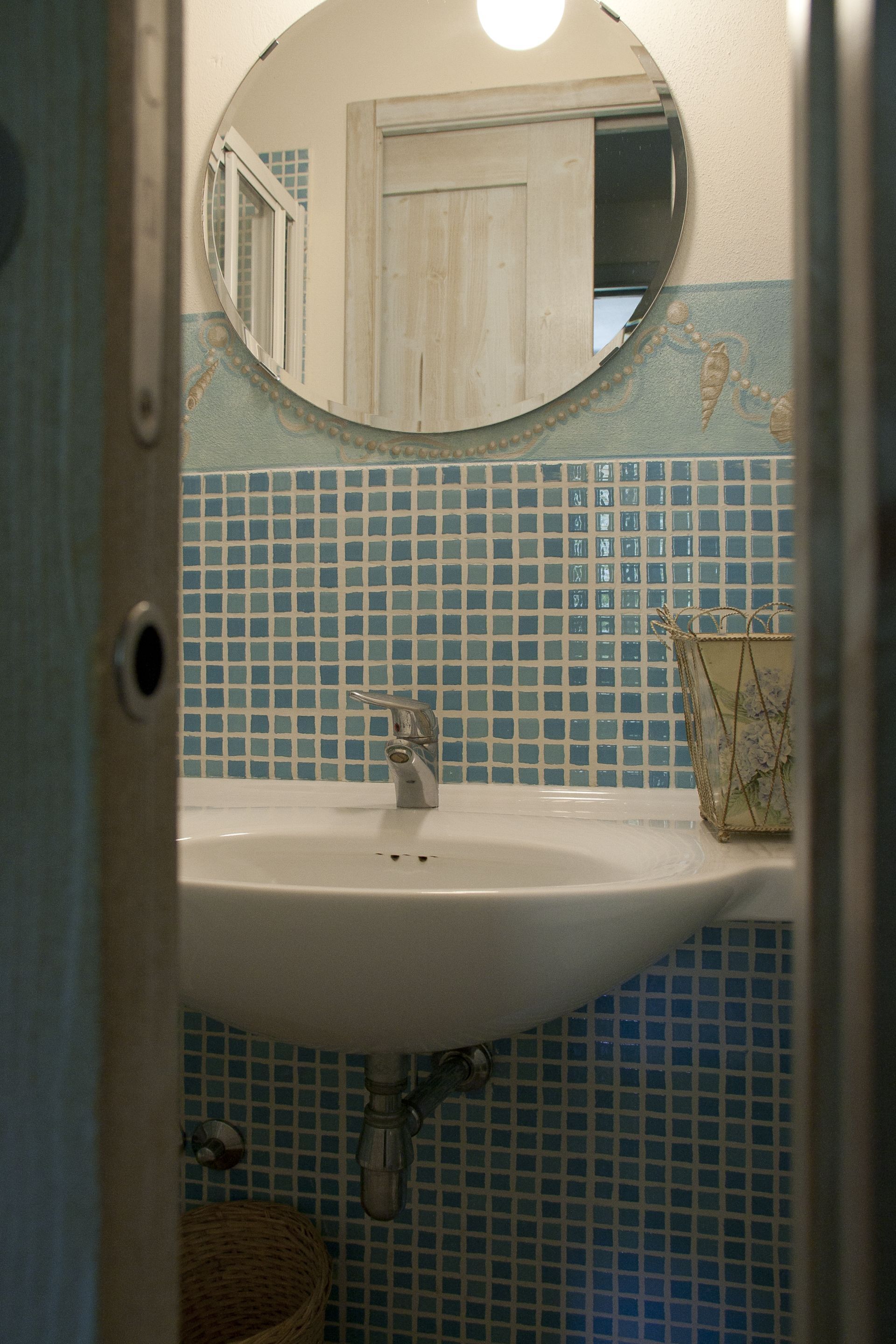 Une salle de bain avec un lavabo blanc, des carreaux de mosaïque bleus, un miroir rond et un panier en osier.