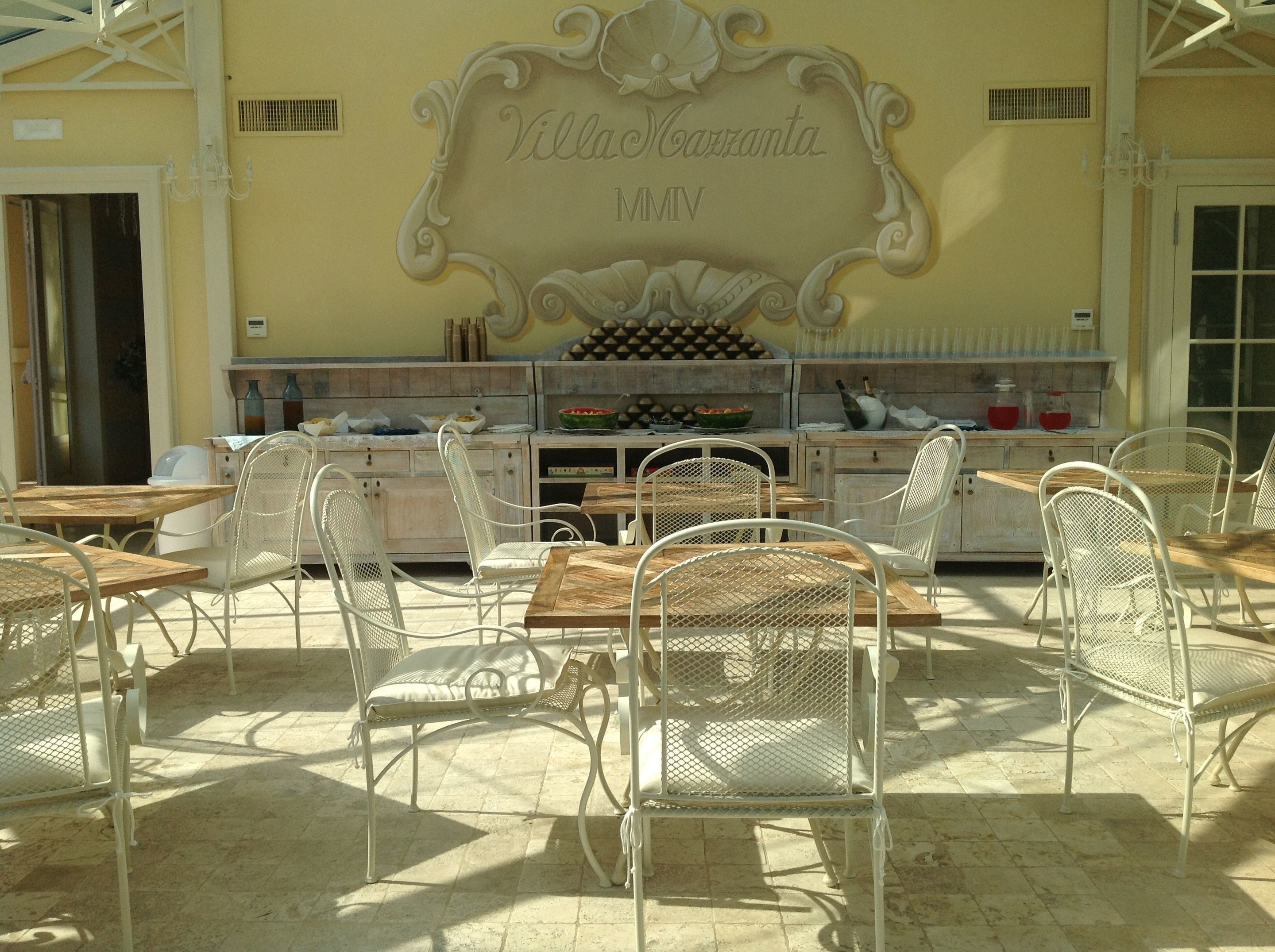 Ein Straßencafé mit weißen Metallstühlen und -tischen, einer Wand mit der Aufschrift "Villa Maranta MMV" und einem Bereich für Speisen und Getränke.