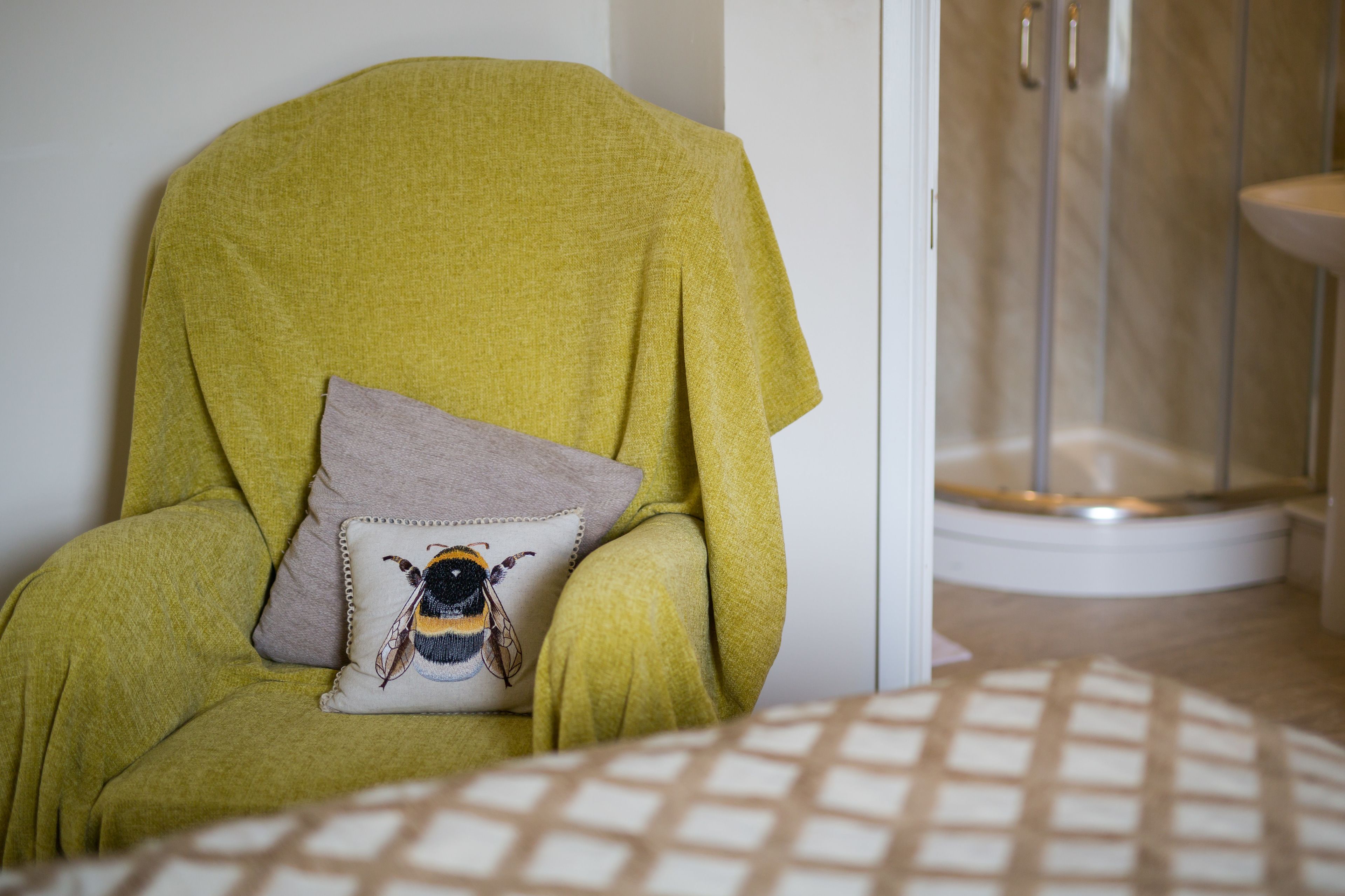 A yellow armchair with a beige pillow featuring a bee design, positioned in a bathroom with a white sink and a shower area.