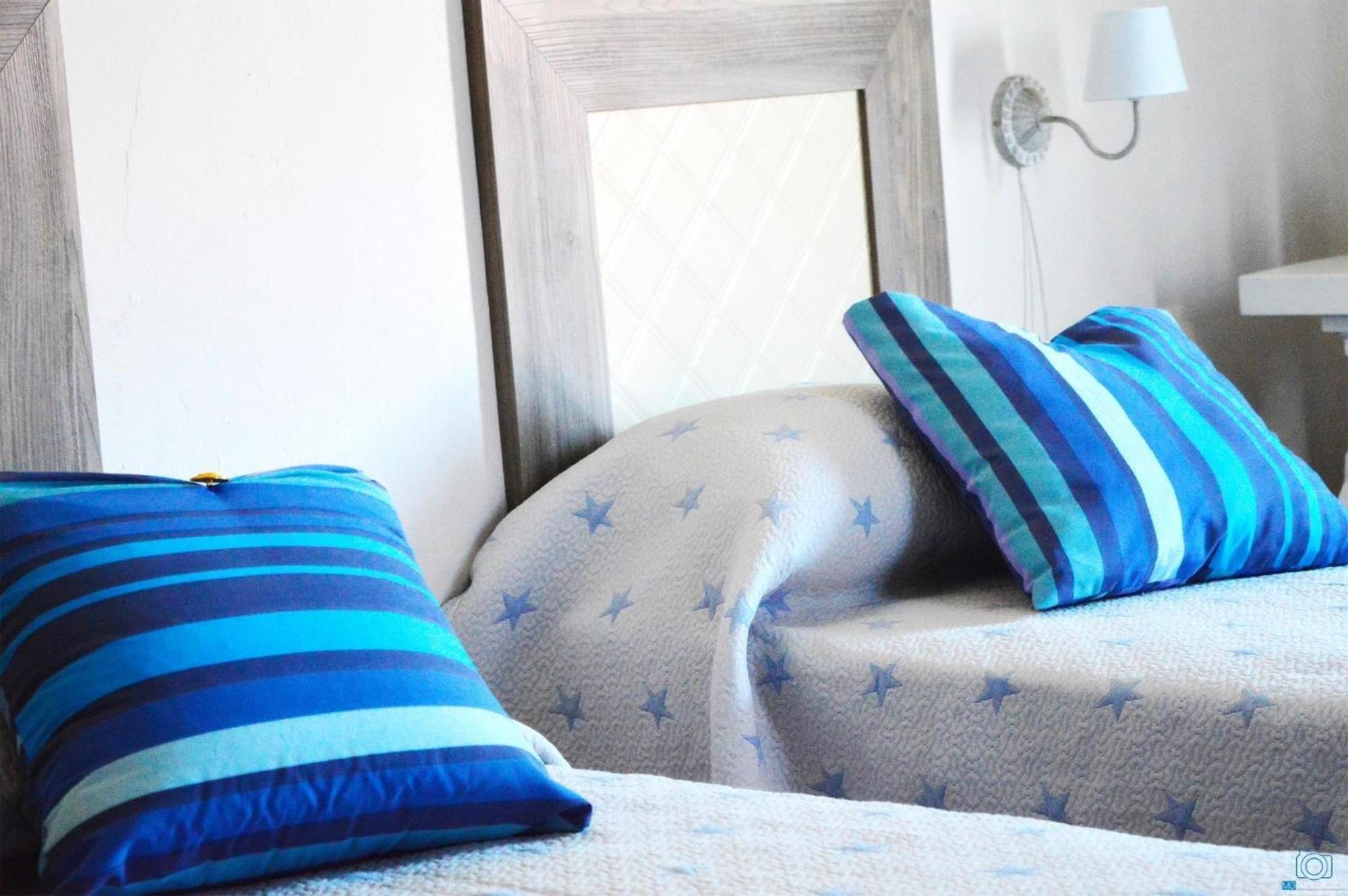 A bedroom with a bed covered in a light-colored duvet with star patterns, two pillows with blue and white stripes, and a wall-mounted lamp.