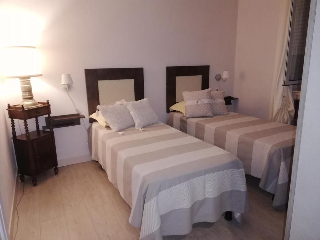 A hotel room with two beds, a bedside table with a lamp, and a window with curtains.