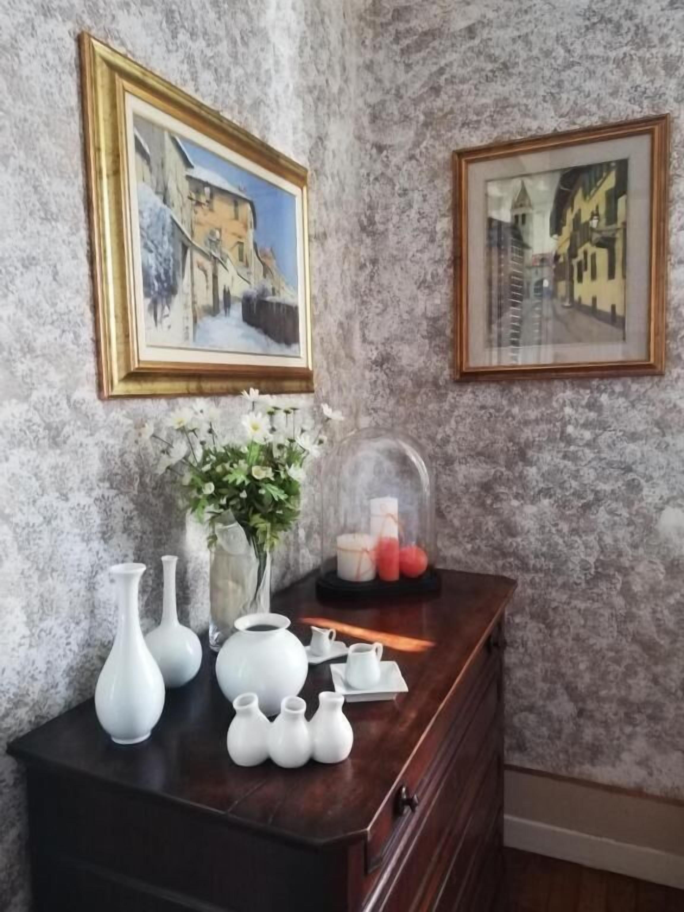 A room with a marble wall, a dark wooden dresser, white vases, a glass dome, and framed paintings.