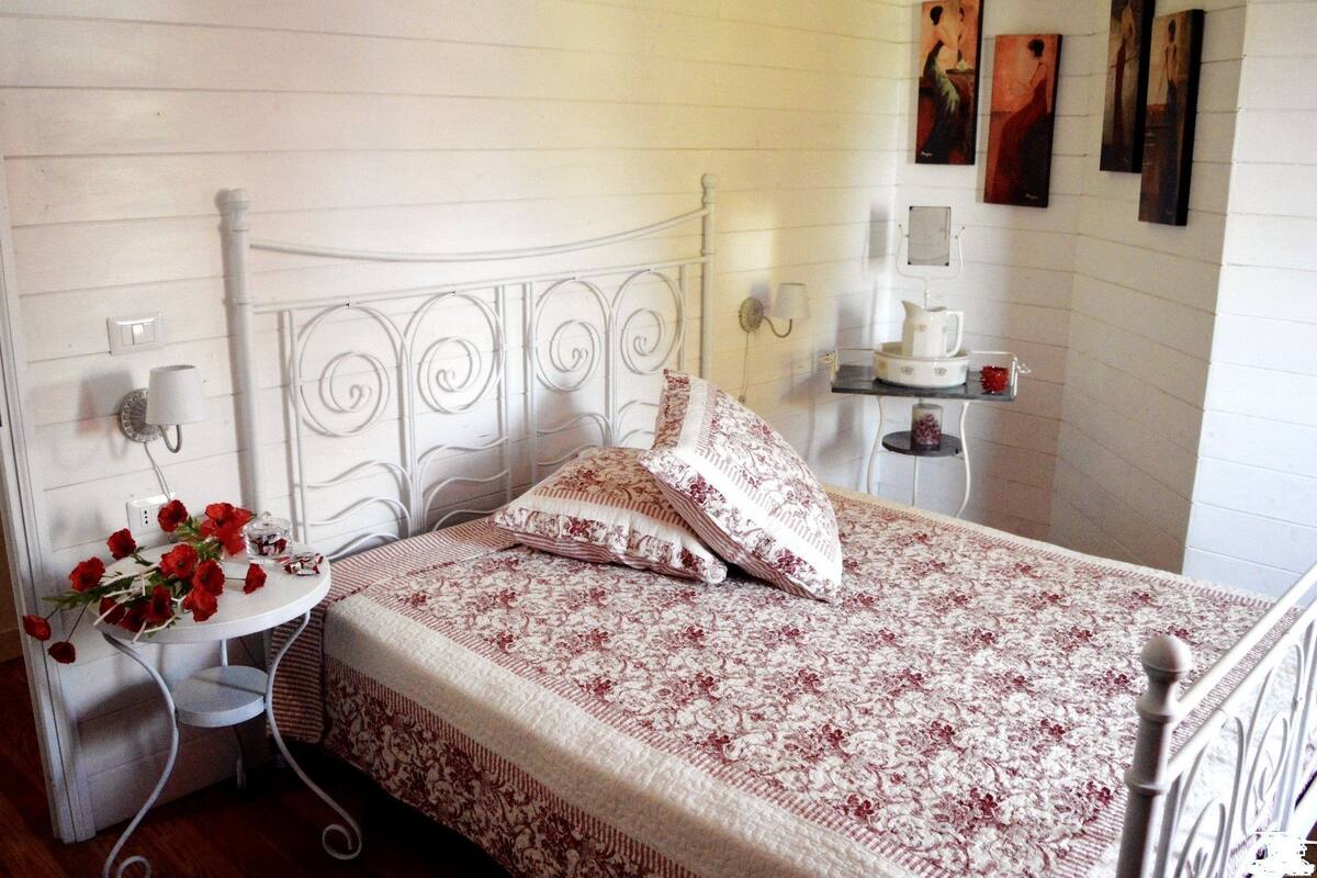 A bedroom with a white metal bed frame, a bedside table with a lamp, and a small shelf with decorative items.