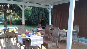 An outdoor patio with wicker chairs and tables, covered by a white pergola, surrounded by greenery.